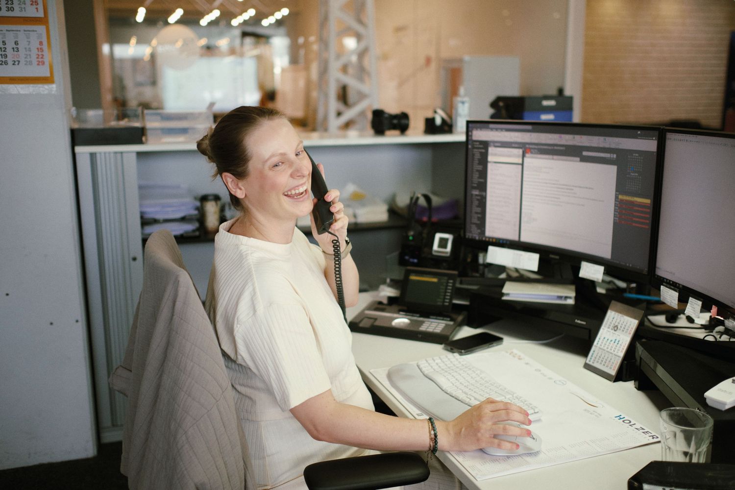Woman smiling while talking on phone at office desk with multiple computer monitors in bright workspace.