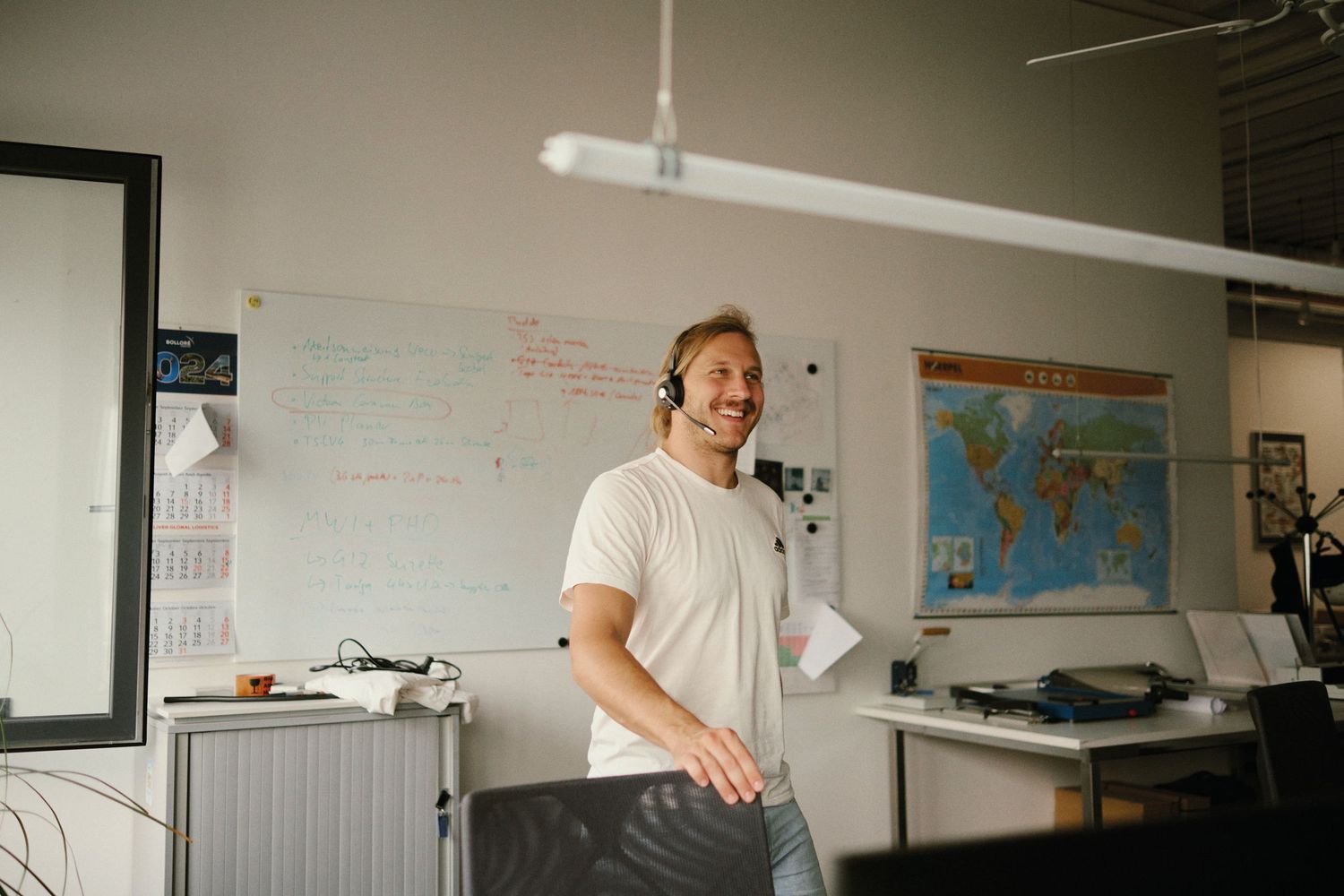 Smiling man wearing headset in modern office with whiteboard and world map on wall.