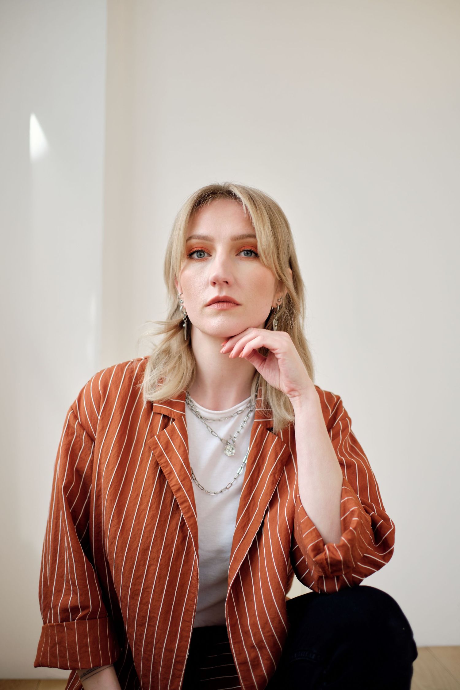 Blonde woman in striped rust blazer and silver necklace posing thoughtfully against a white wall.