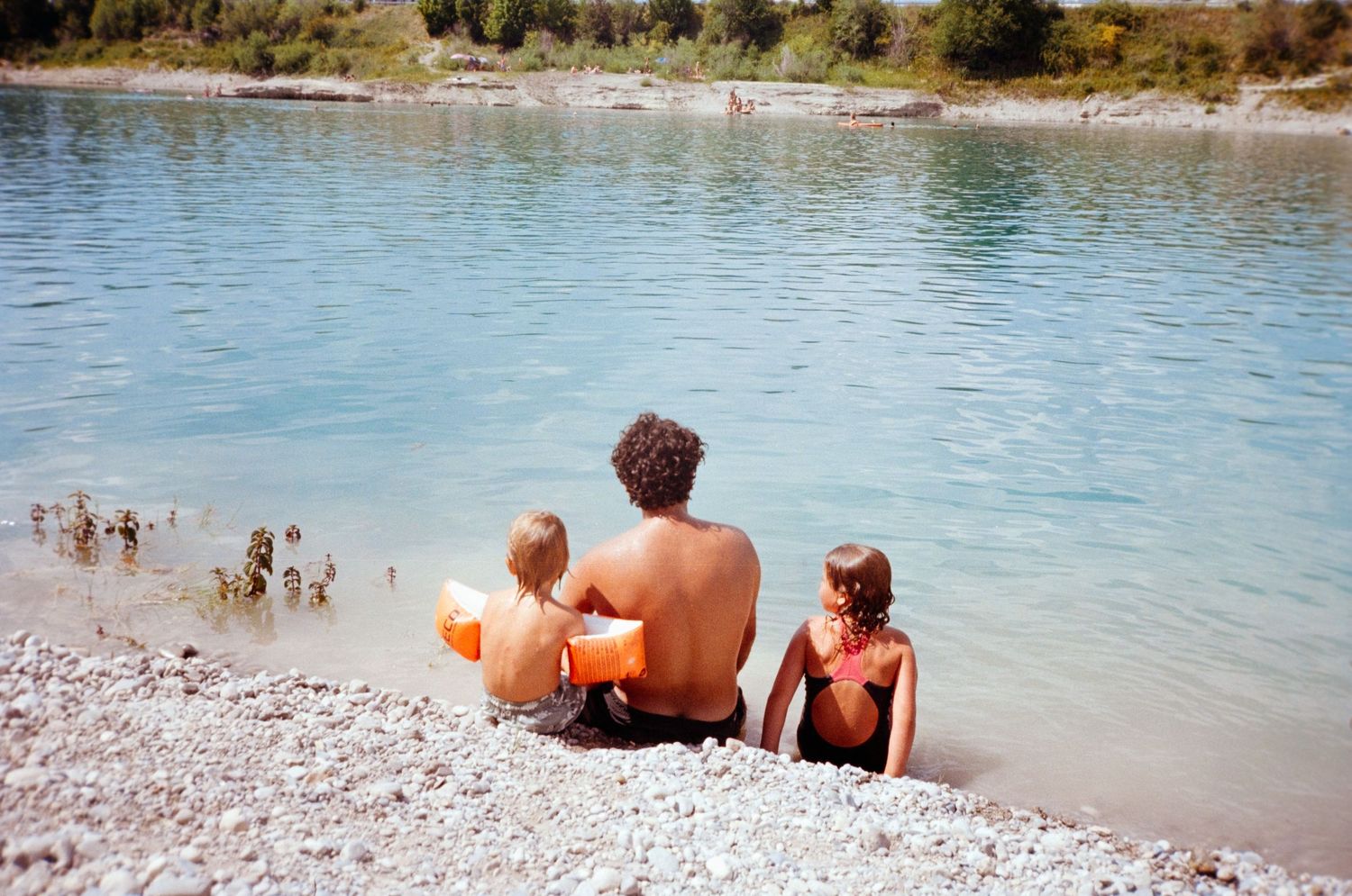 Man and two children sitting on rocky shore by calm turquoise lake on sunny day.