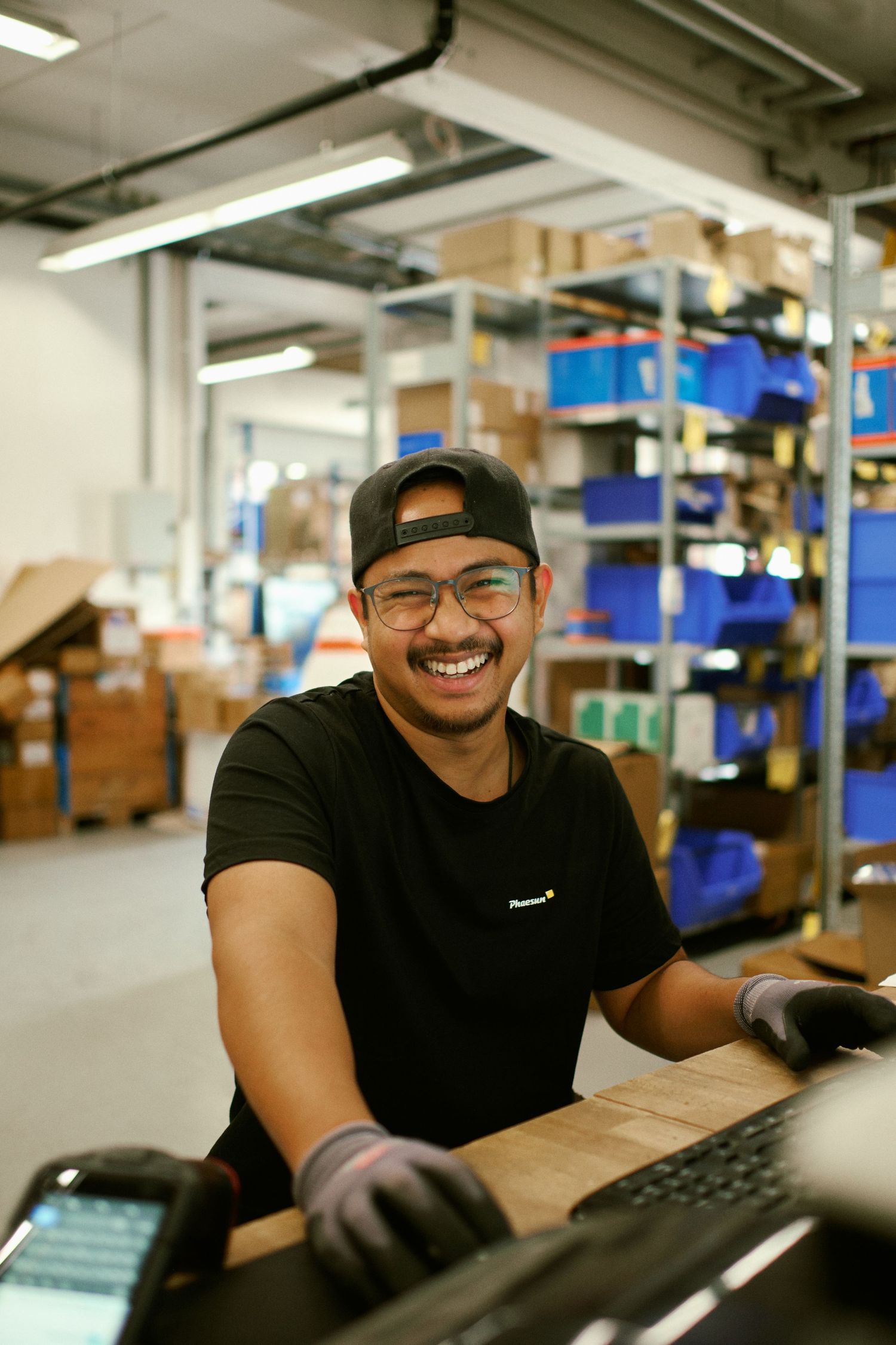 Smiling warehouse worker in black cap and glasses at a workstation surrounded by shelving with boxes.