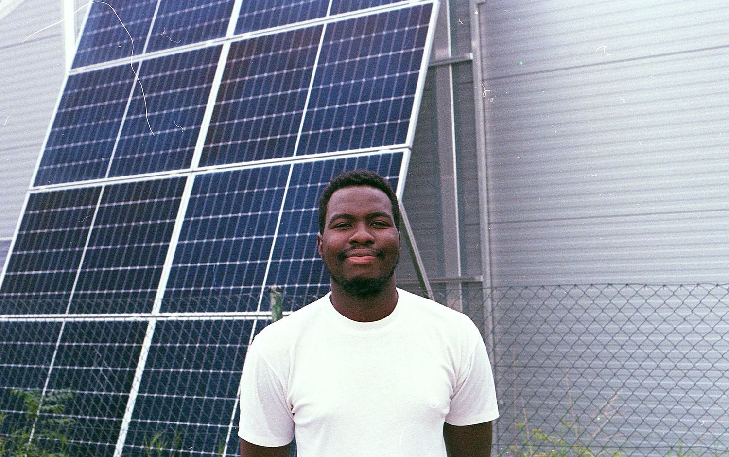 Man in white t-shirt smiling in front of large solar panels installation outdoors.
