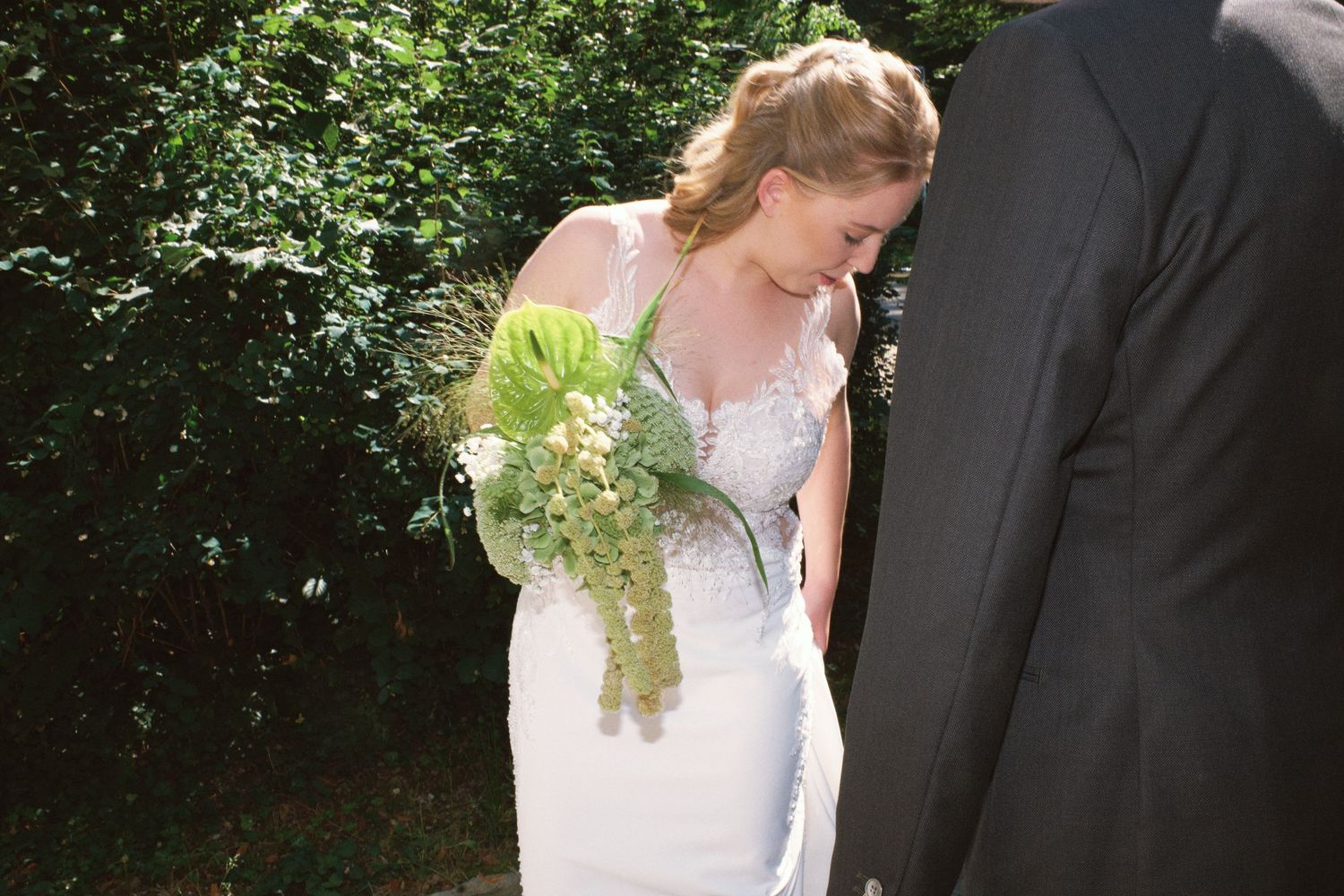 Bride in white lace wedding gown holding green cascading bouquet, standing outdoors near lush greenery.