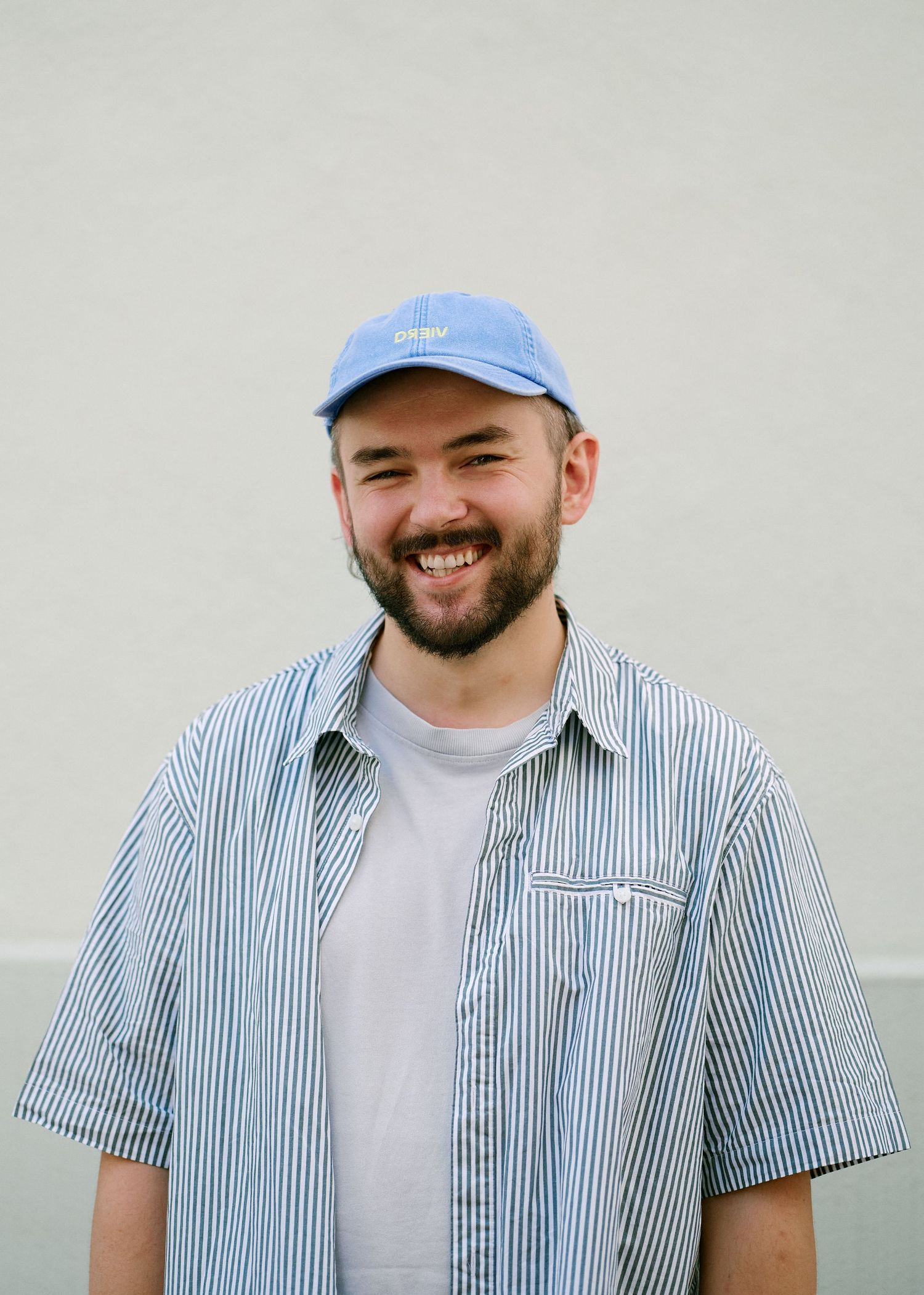 Smiling bearded man wearing a blue striped shirt and blue baseball cap standing against a light gray wall.