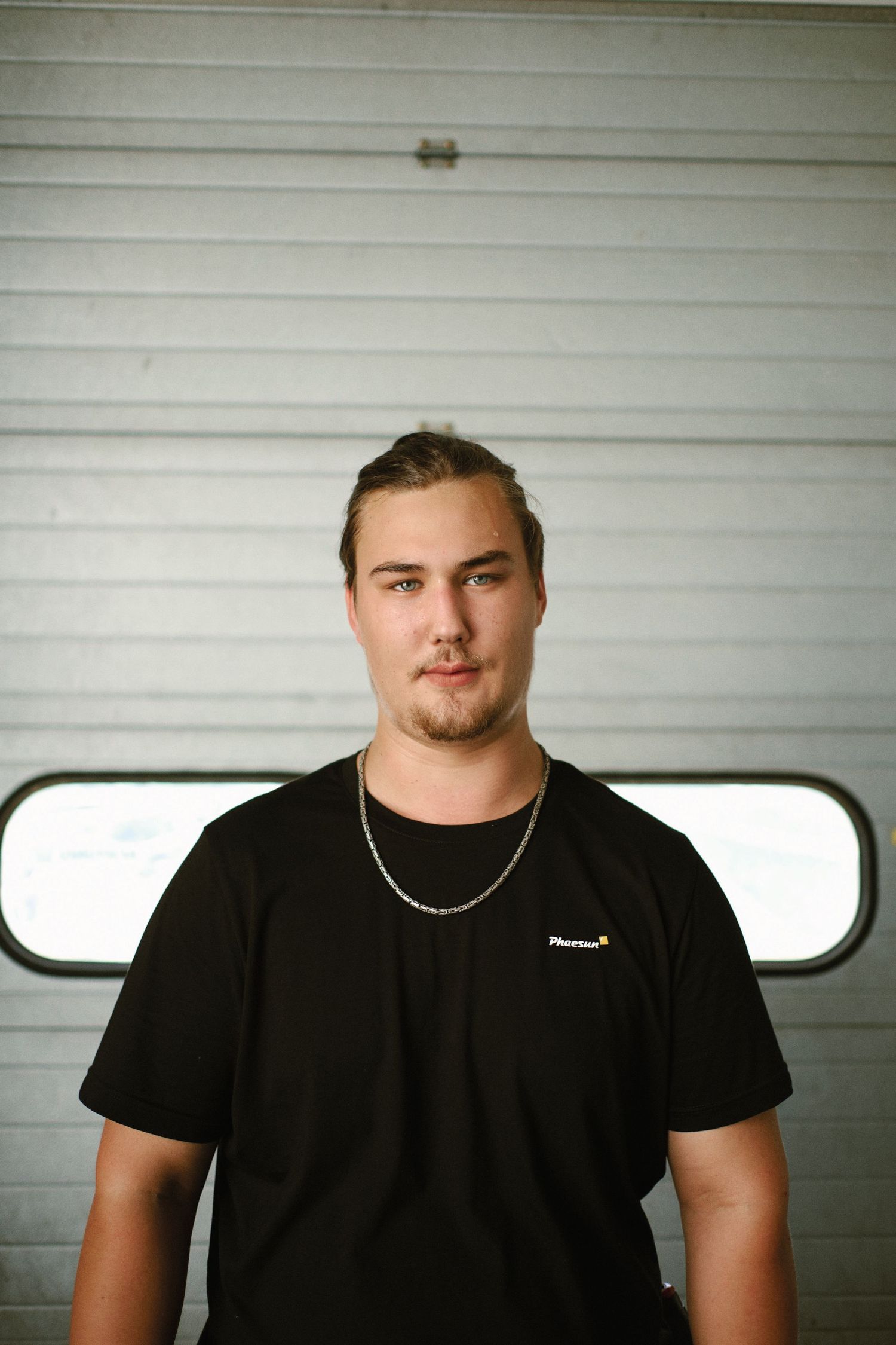 Young man in black t-shirt and chain necklace standing in front of a garage door.
