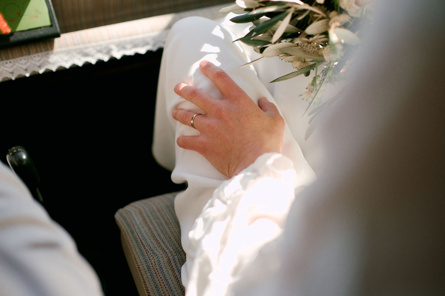 Close-up of a bride's hand wearing a gold wedding ring, holding white fabric with flowers in soft sunlight.