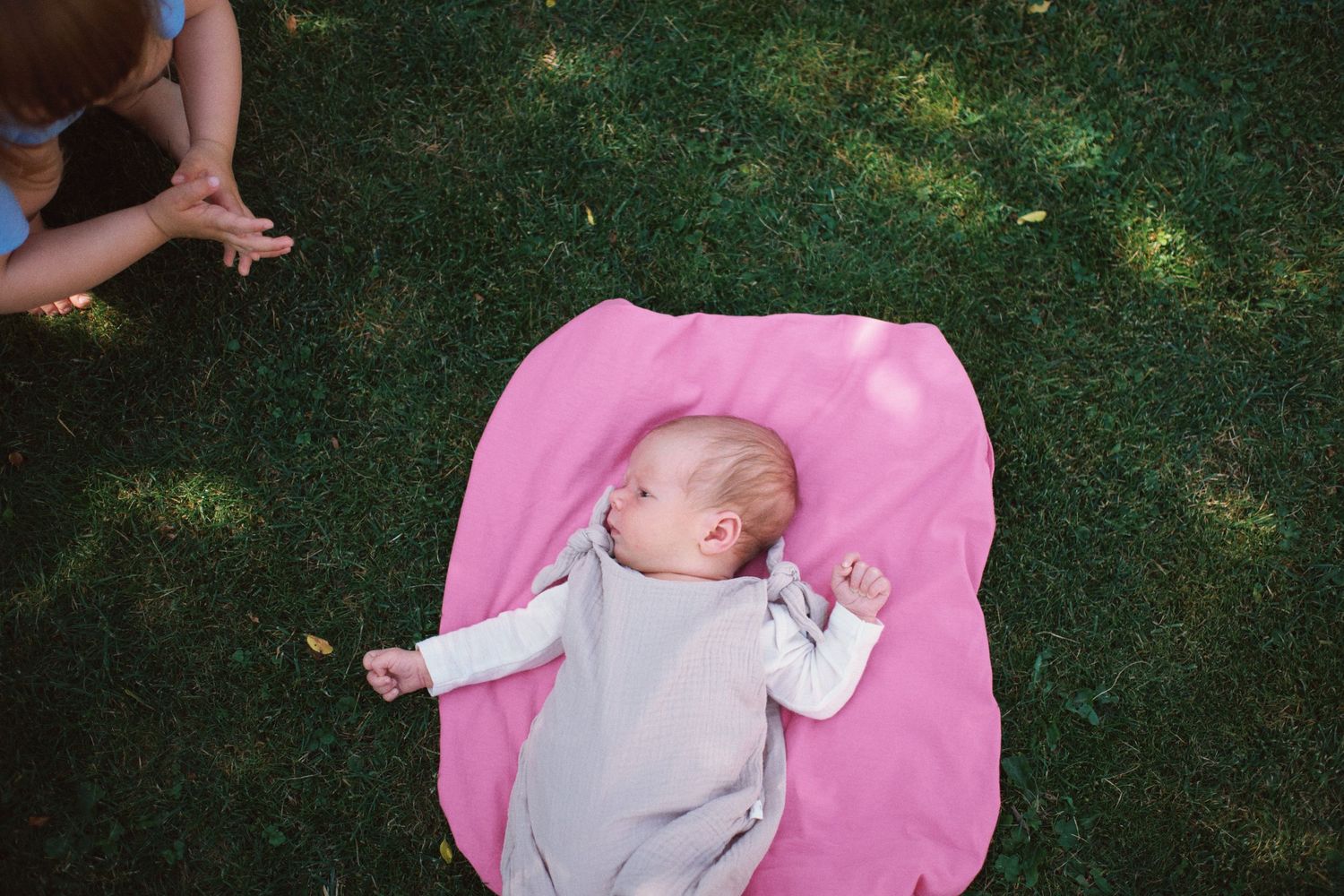Newborn baby lying on a pink blanket on green grass outdoors, wearing a gray swaddle wrap.