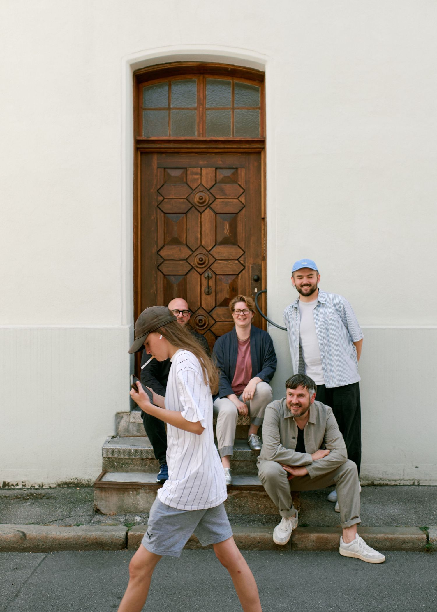 Five young adults posing casually on steps in front of a ornate wooden door on a white building.