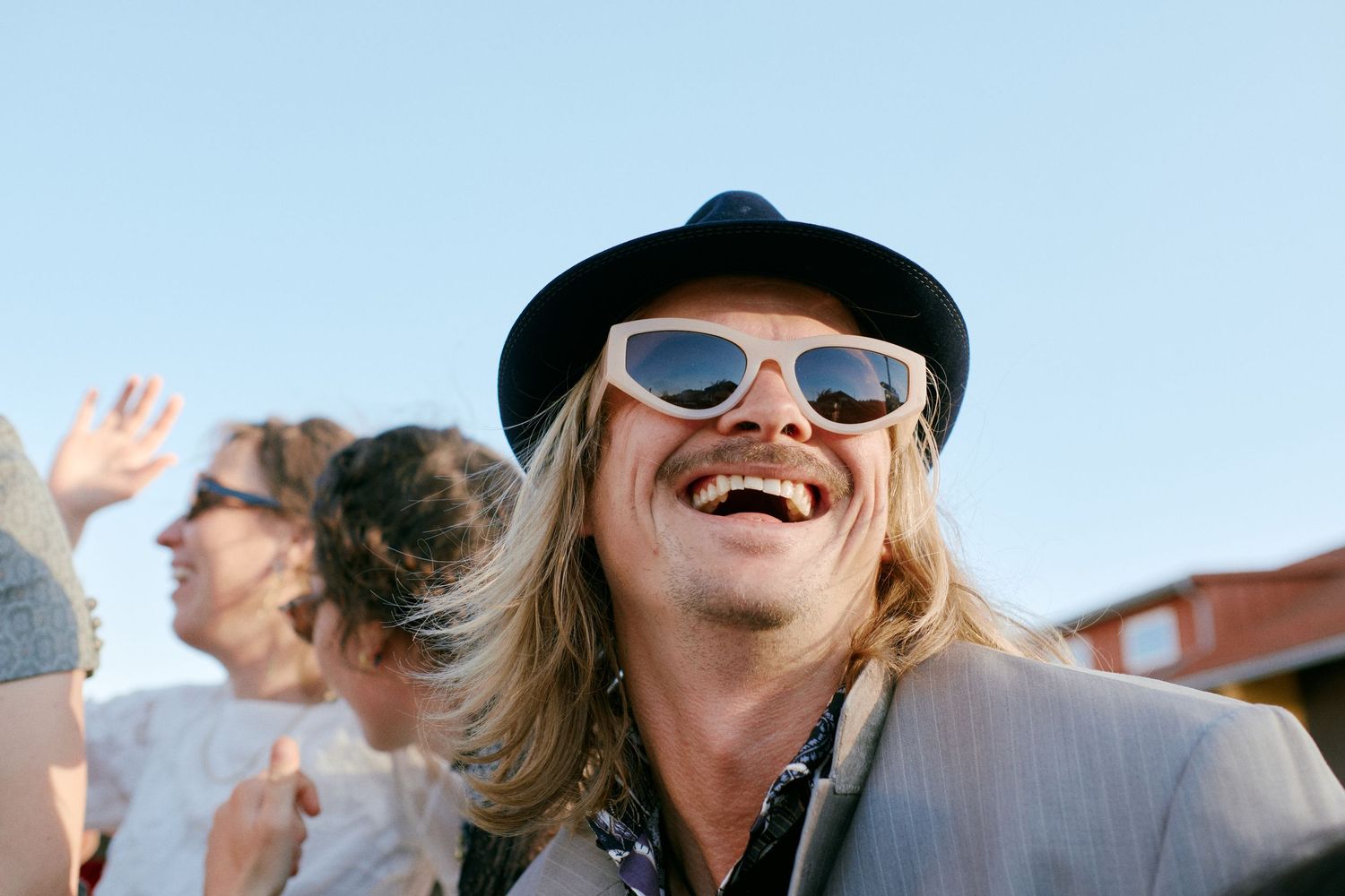 Laughing man with long blonde hair, black hat, and white sunglasses enjoying an outdoor event on a sunny day.
