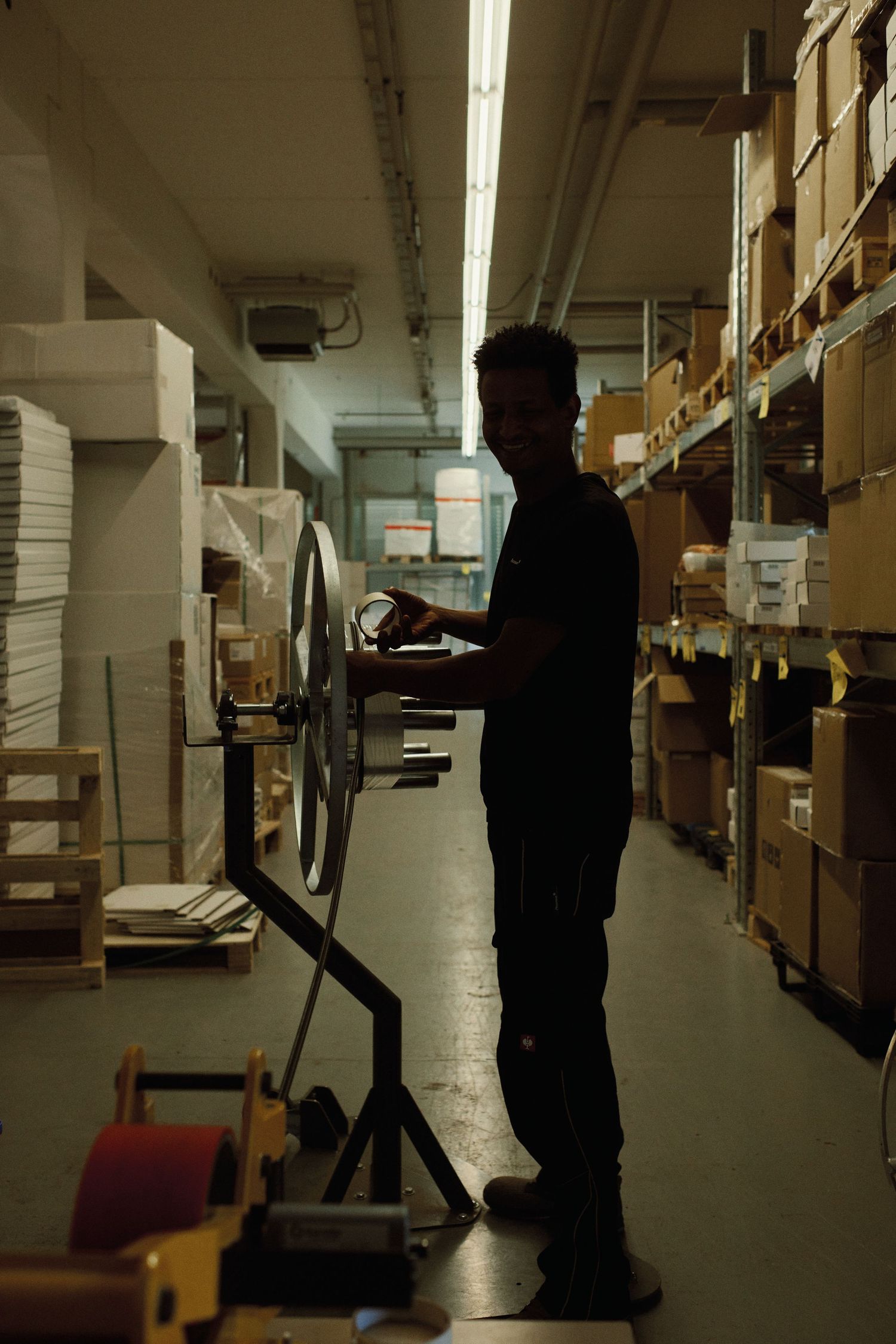 A worker operates machinery in a dimly lit warehouse aisle surrounded by shelves of boxes.