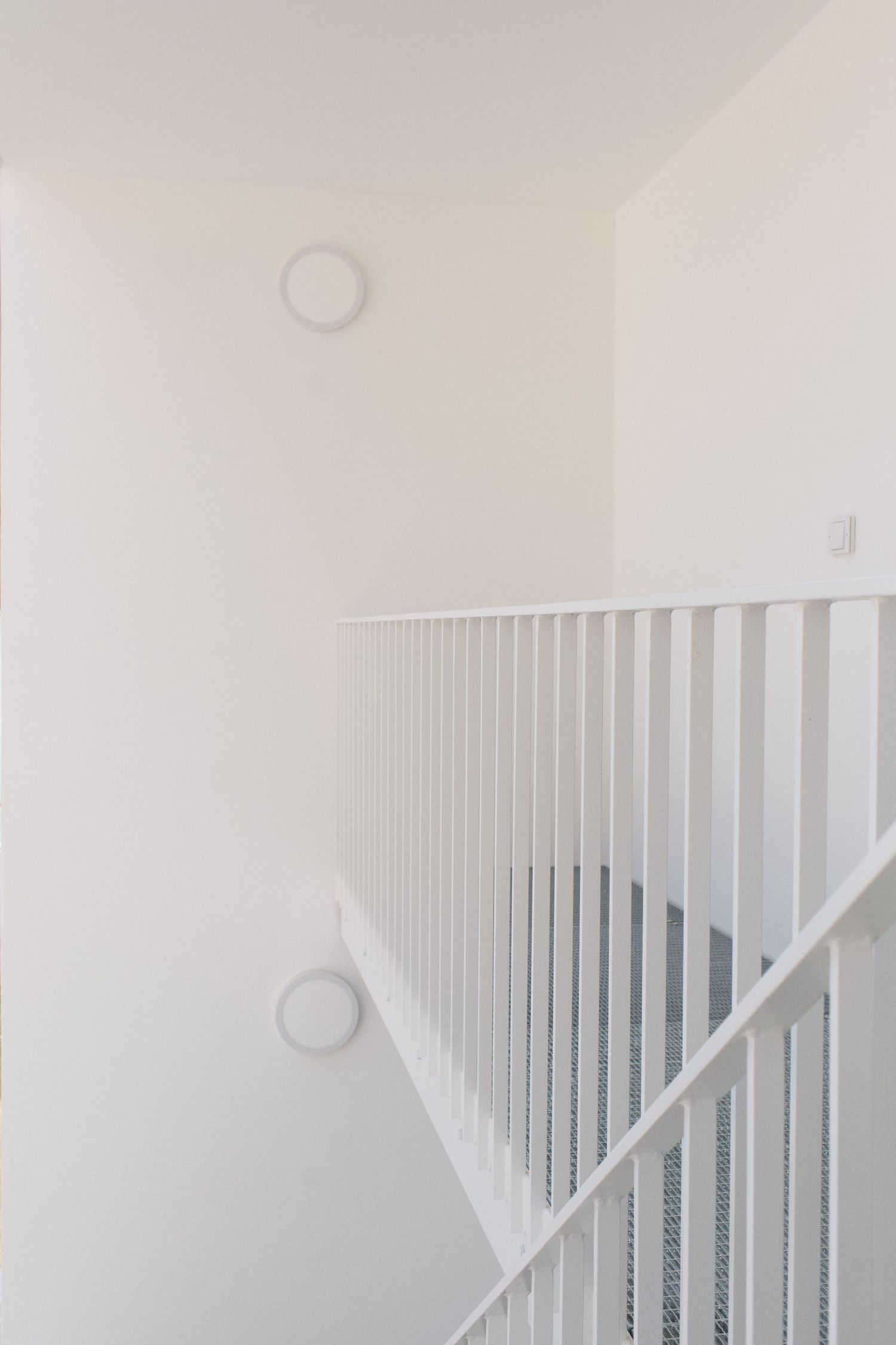 Minimalist white staircase with white balusters and handrail, bright walls, and round wall lights in a modern interior.