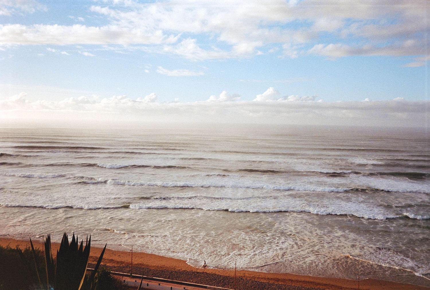 Panoramic ocean view with rolling waves under a partly cloudy sky above a sandy beach coastline.