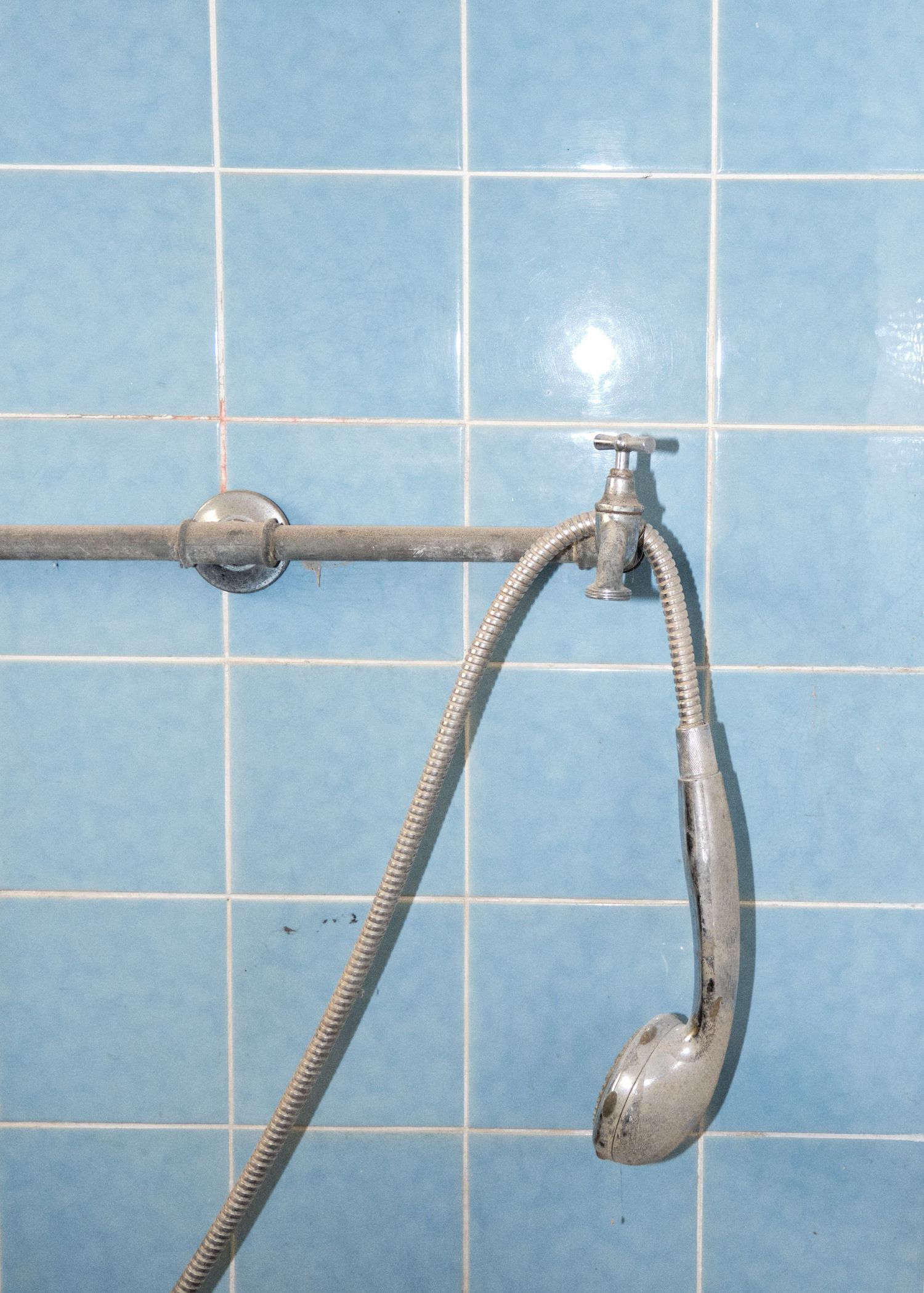 A chrome handheld shower head with flexible hose attached to a wall faucet against blue ceramic tiles.