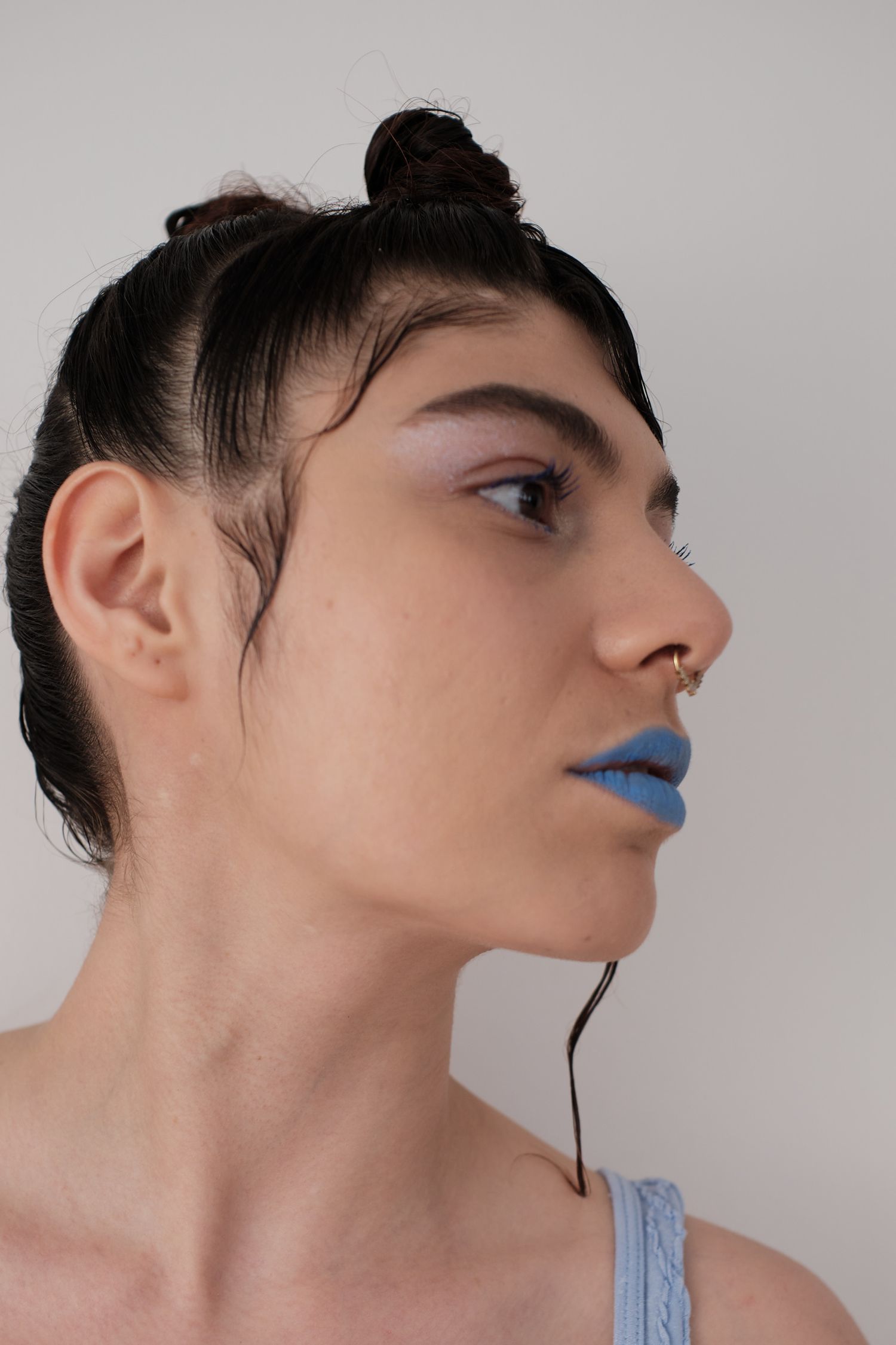 Profile view of a person with blue matte lipstick, nose ring, hair bun, and subtle eye makeup against white background.