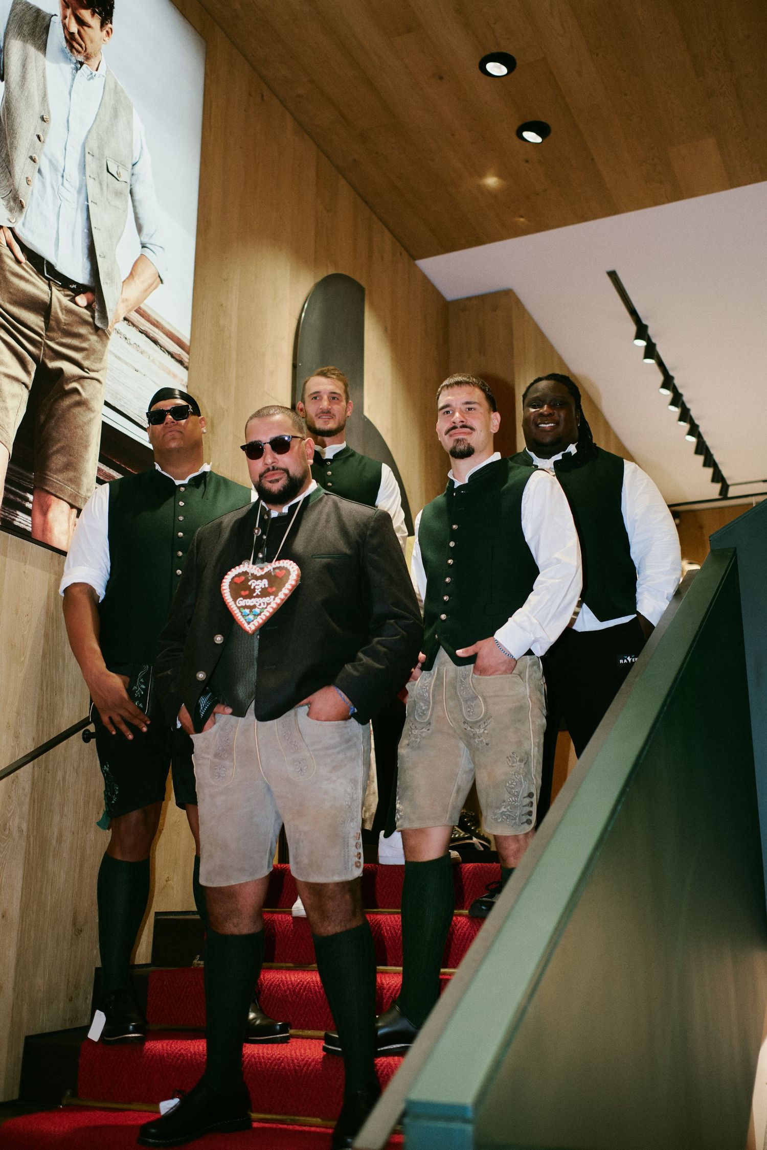 Five men in traditional Bavarian lederhosen and vests pose on red-carpeted stairs in a modern interior.