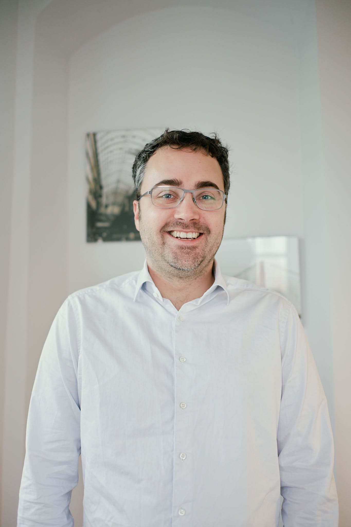 Smiling man with glasses and dark curly hair wearing a white button-up shirt standing in a bright modern office.