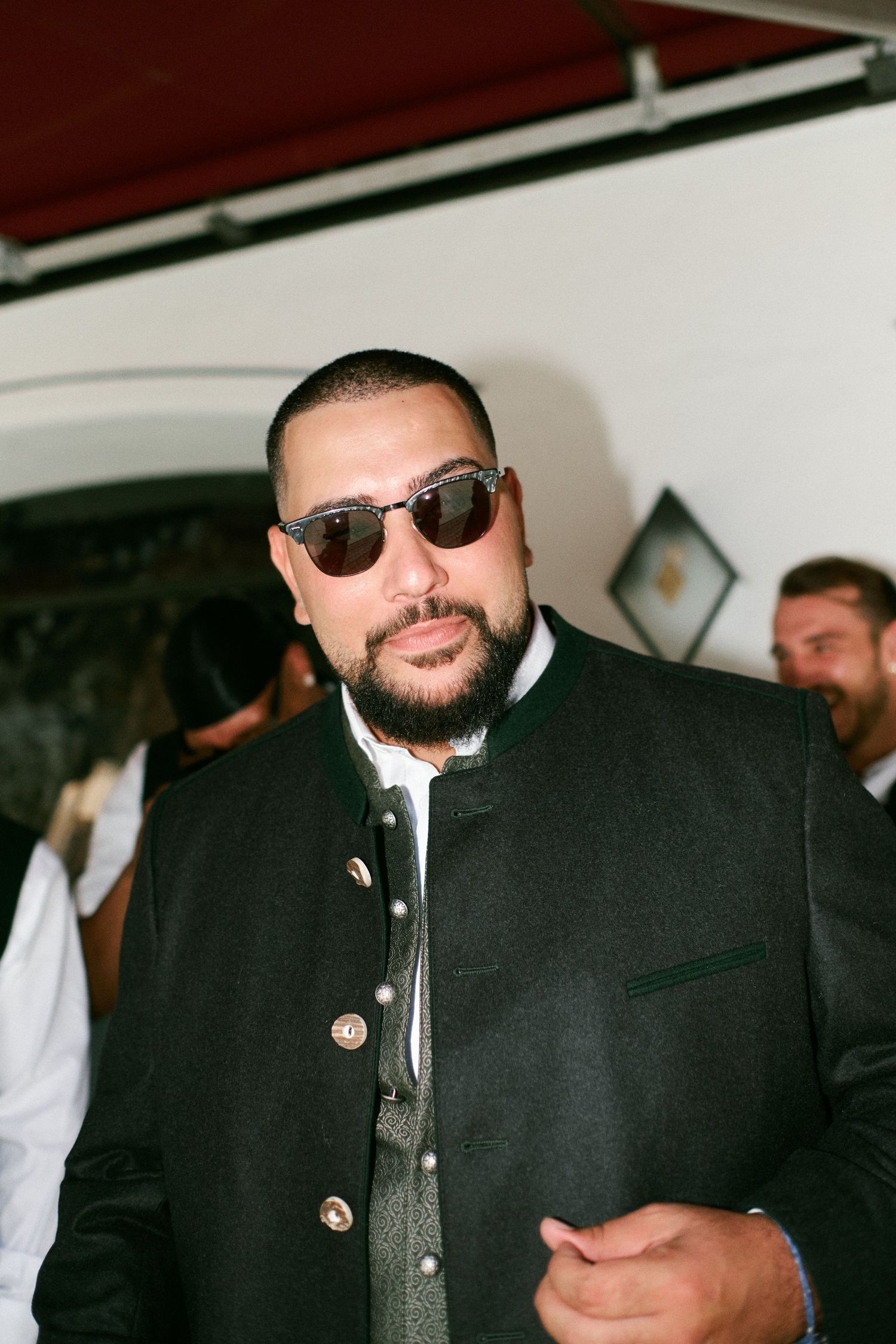 Man with beard and sunglasses wearing a dark traditional jacket at an indoor event.