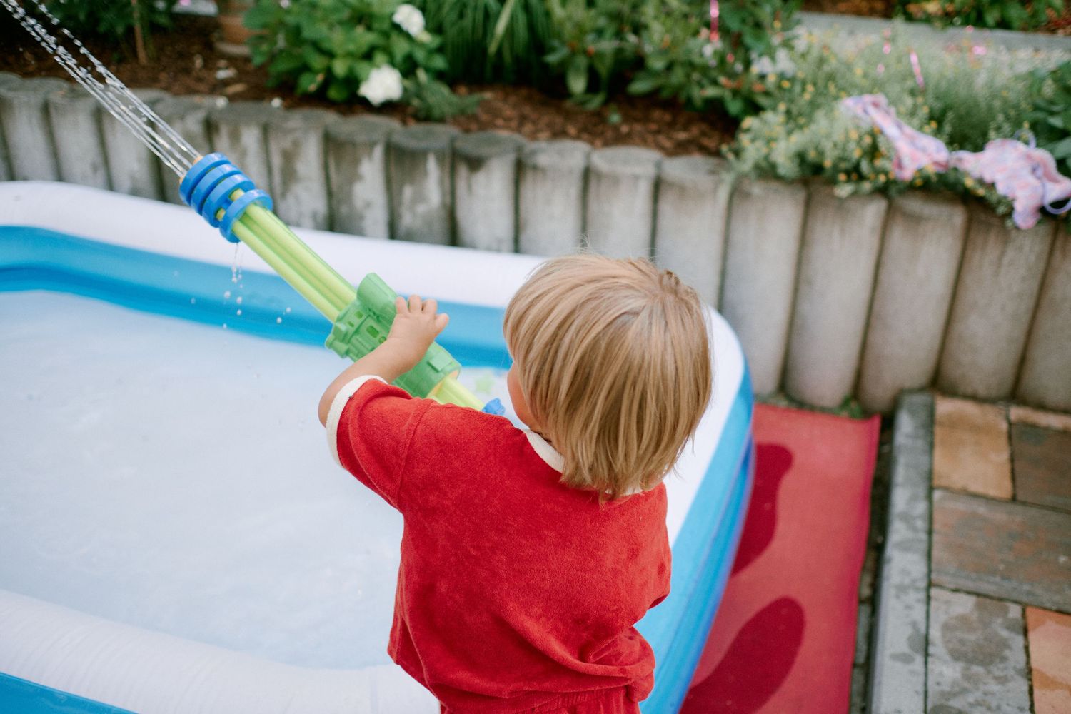 Young toddler in red shirt filling an inflatable backyard pool with a green water toy on a sunny day.