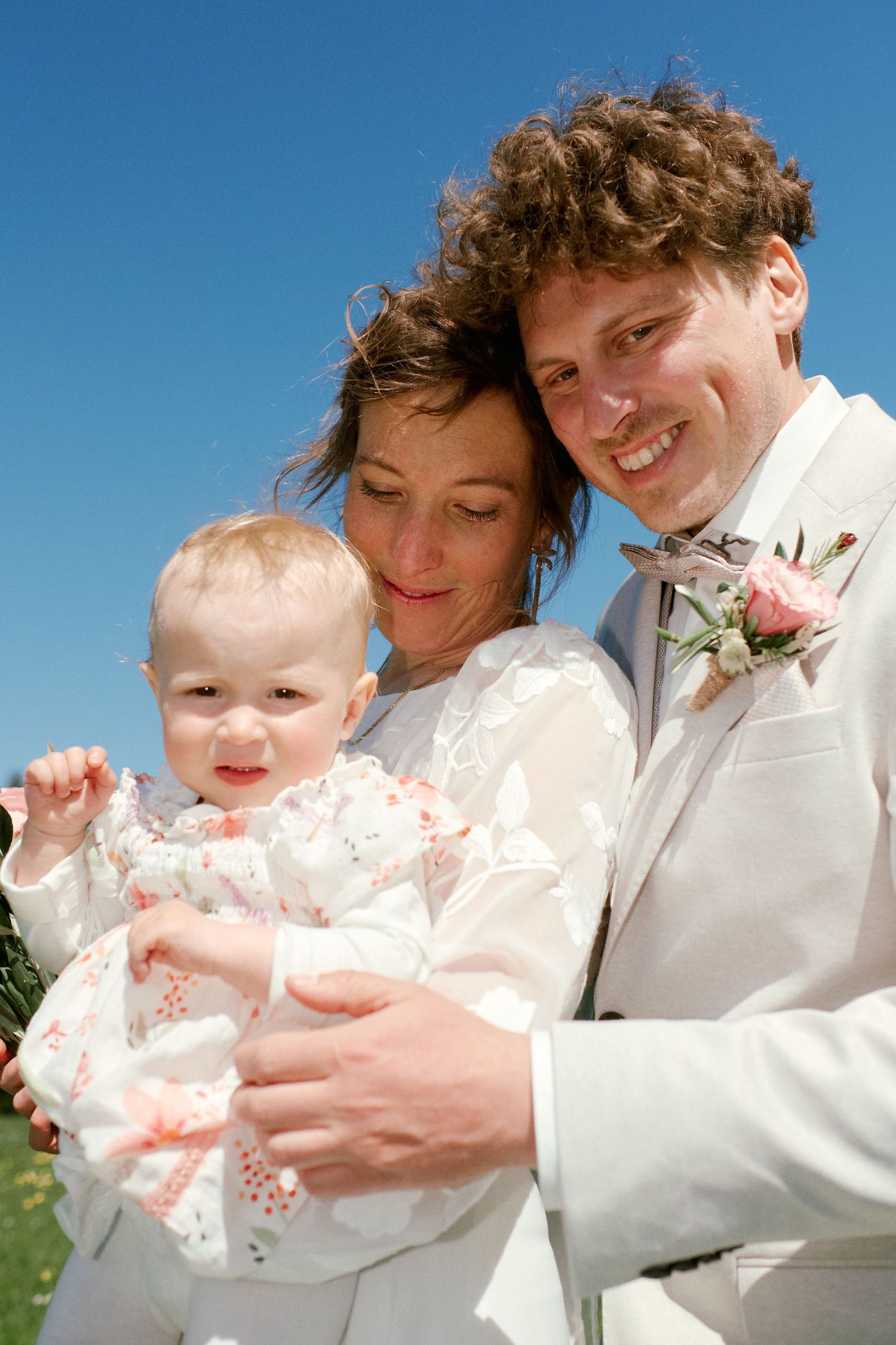 Happy newlywed couple in white wedding attire smiling outdoors, holding their baby girl under a bright blue sky.