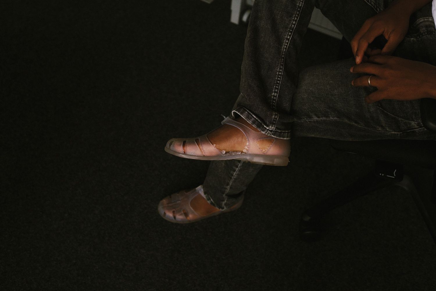 Person sitting cross-legged wearing brown leather shoes and gray jeans in a dimly lit setting.