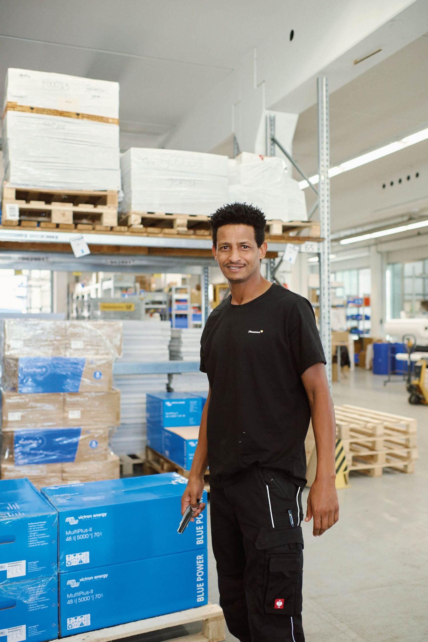 A smiling warehouse worker in black clothing stands among shelves stocked with blue boxes and pallets in a storage facility.