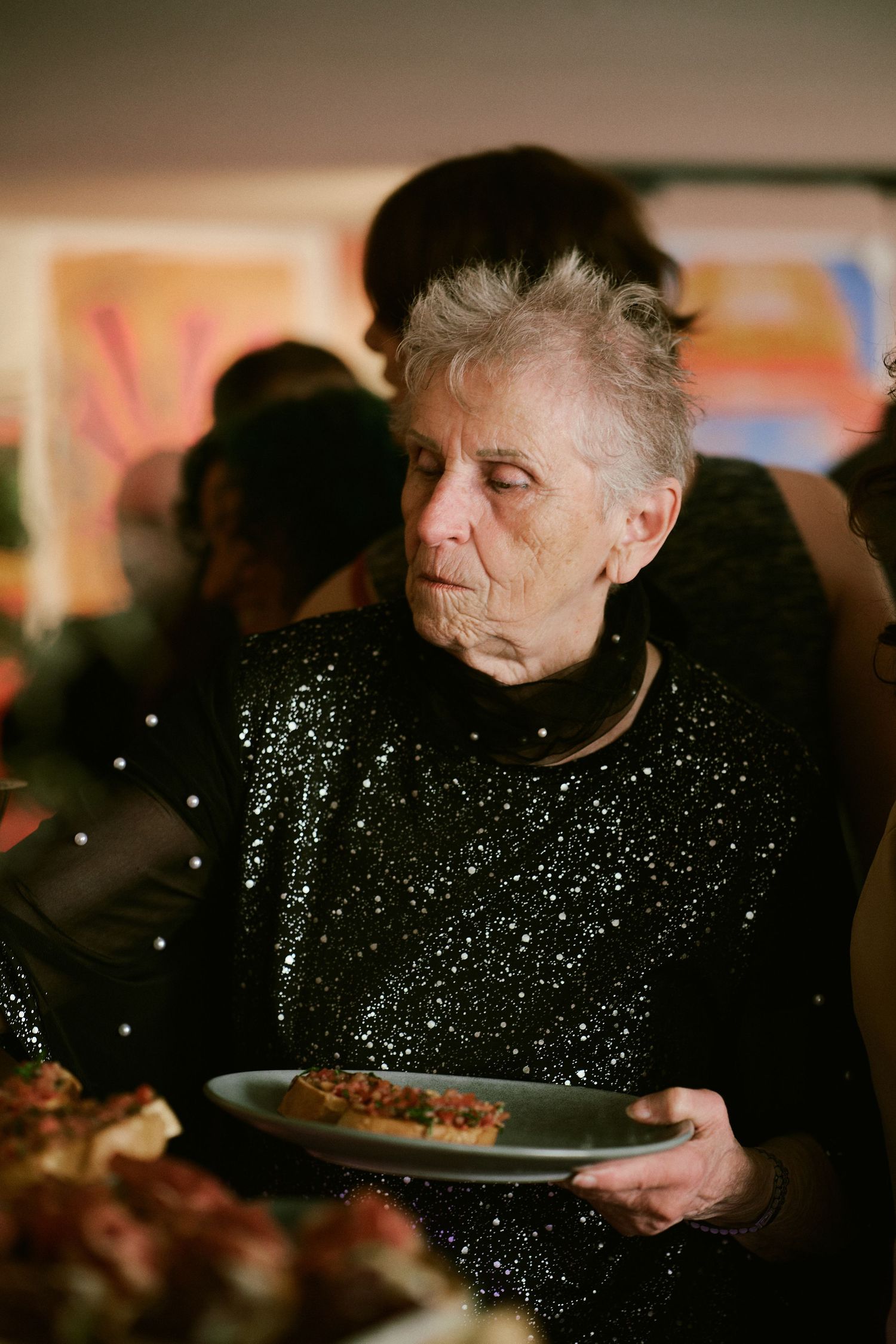 Elderly woman in black sequin top holds plate at social gathering with colorful artwork in background.