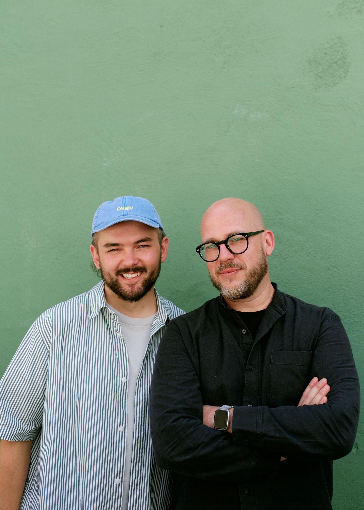 Two men smiling against a green wall, one wearing a blue cap and striped shirt, the other bald with glasses and black shirt.