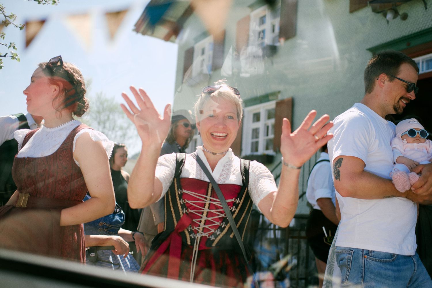 Woman in traditional dirndl dress waving and smiling at outdoor festival with crowd in background.