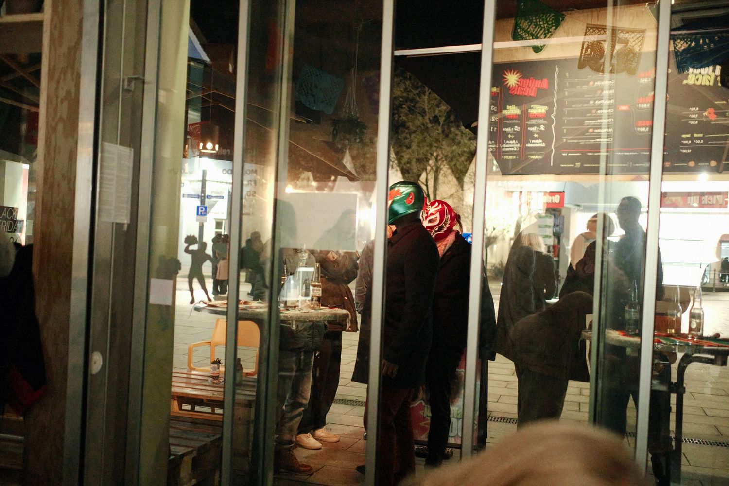 People wearing masks standing inside a store entrance at night, with reflections visible in glass doors.