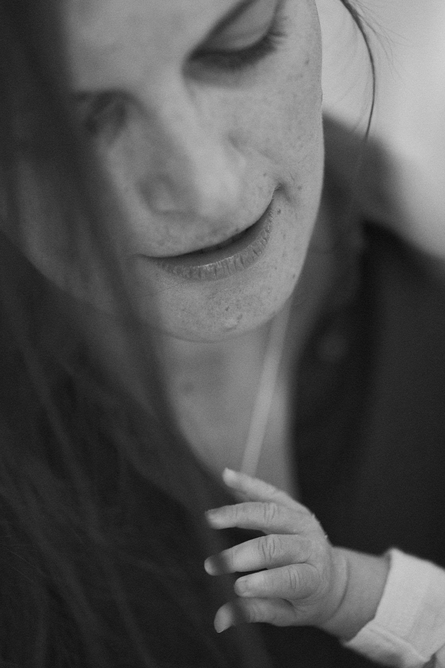 Black and white portrait of a smiling woman looking down at a newborn baby's tiny hand touching her chin.