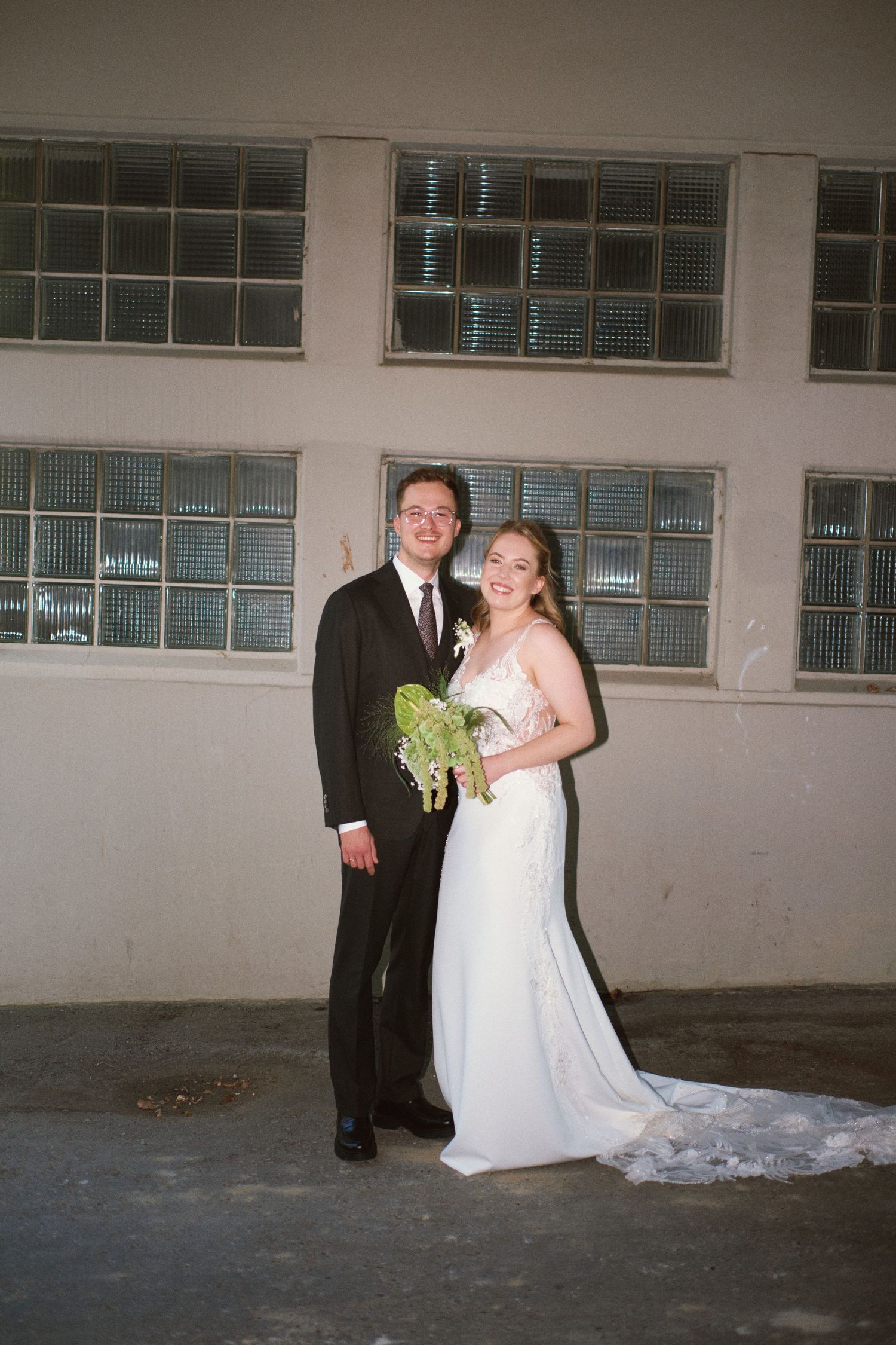 Bride and groom posing together in white dress and black suit, holding green bouquet in industrial venue.