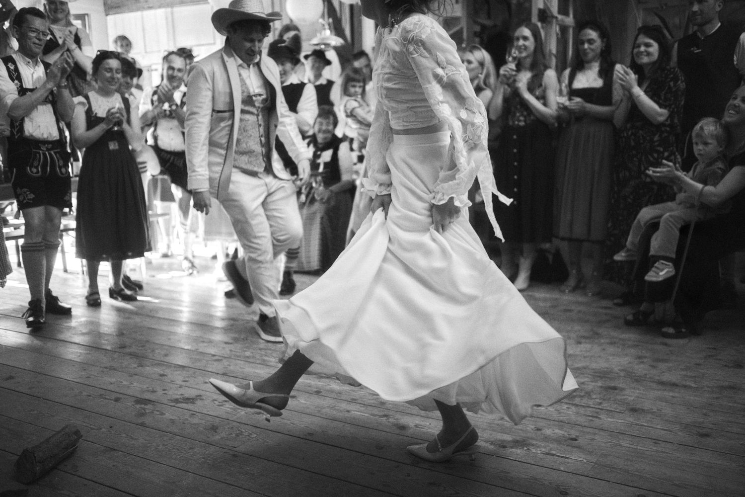 Black and white photo of dancers in traditional folk costumes performing energetically on a wooden dance floor at a cultural event.