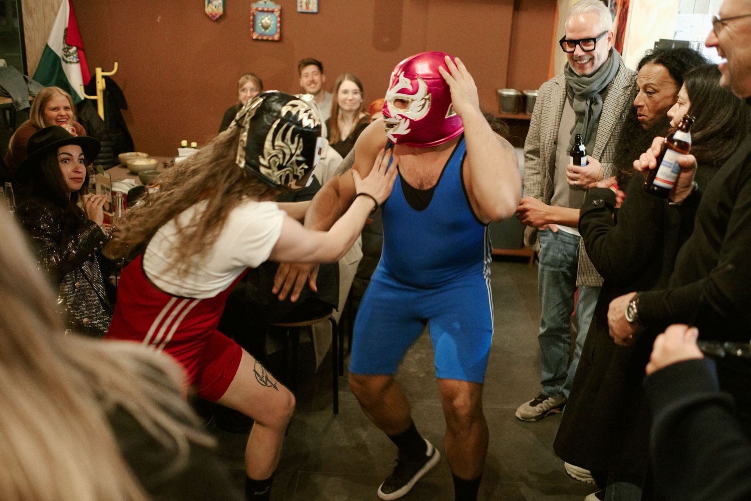 Two people in colorful lucha libre wrestling masks perform for a crowd at an indoor party gathering.