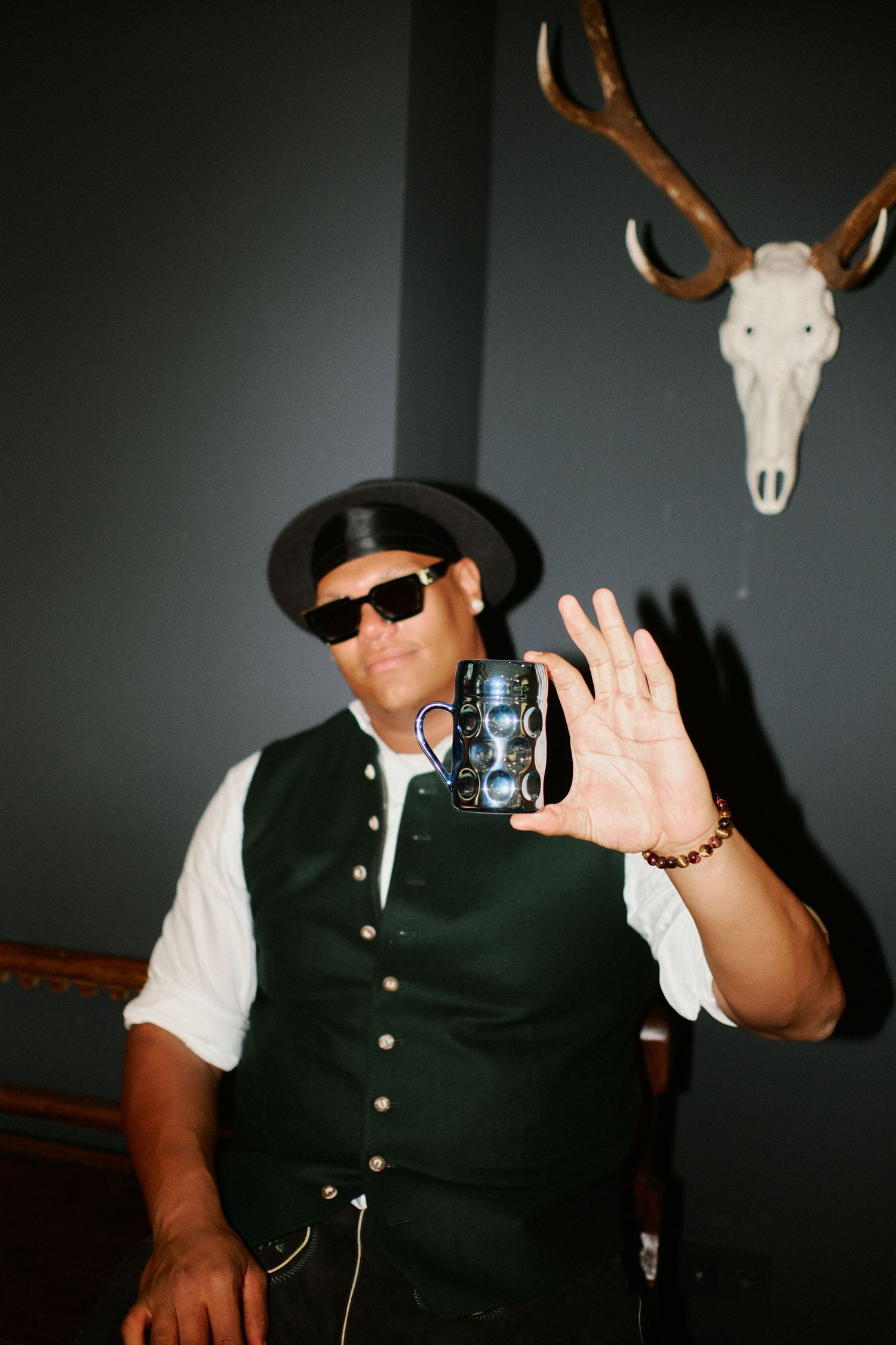 Man in black hat and sunglasses holds vintage camera while posing near mounted deer skull on gray wall.