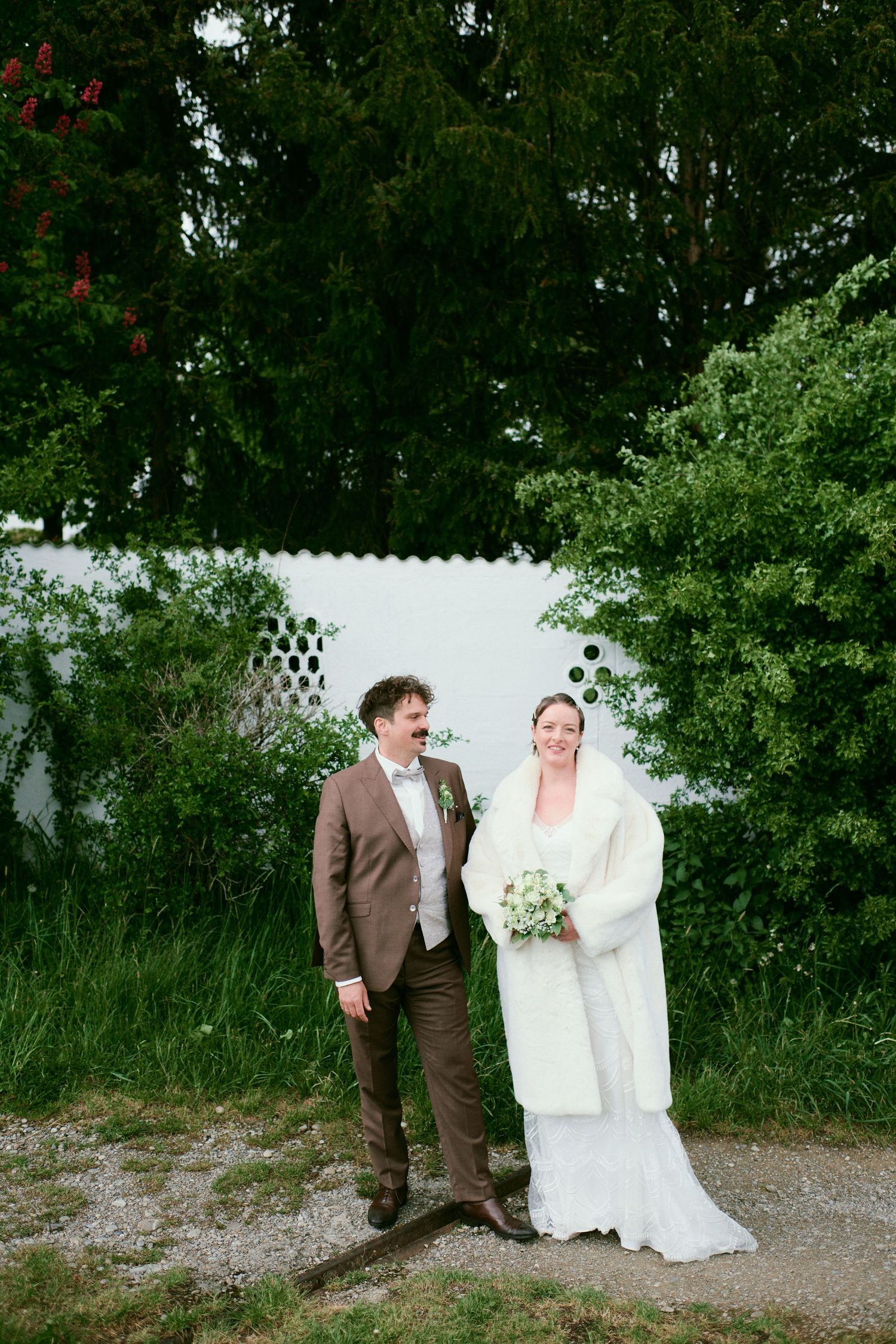 Bride in white gown and fur wrap holds bouquet beside groom in brown suit, standing outdoors near green trees.