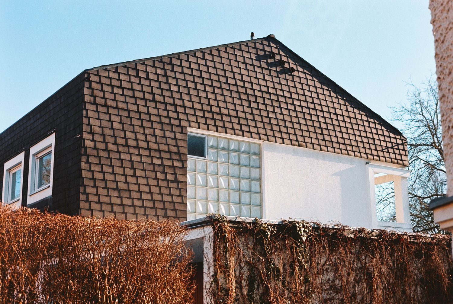 Dark tile-roofed house with glass block window feature, surrounded by autumn hedges under clear blue sky.