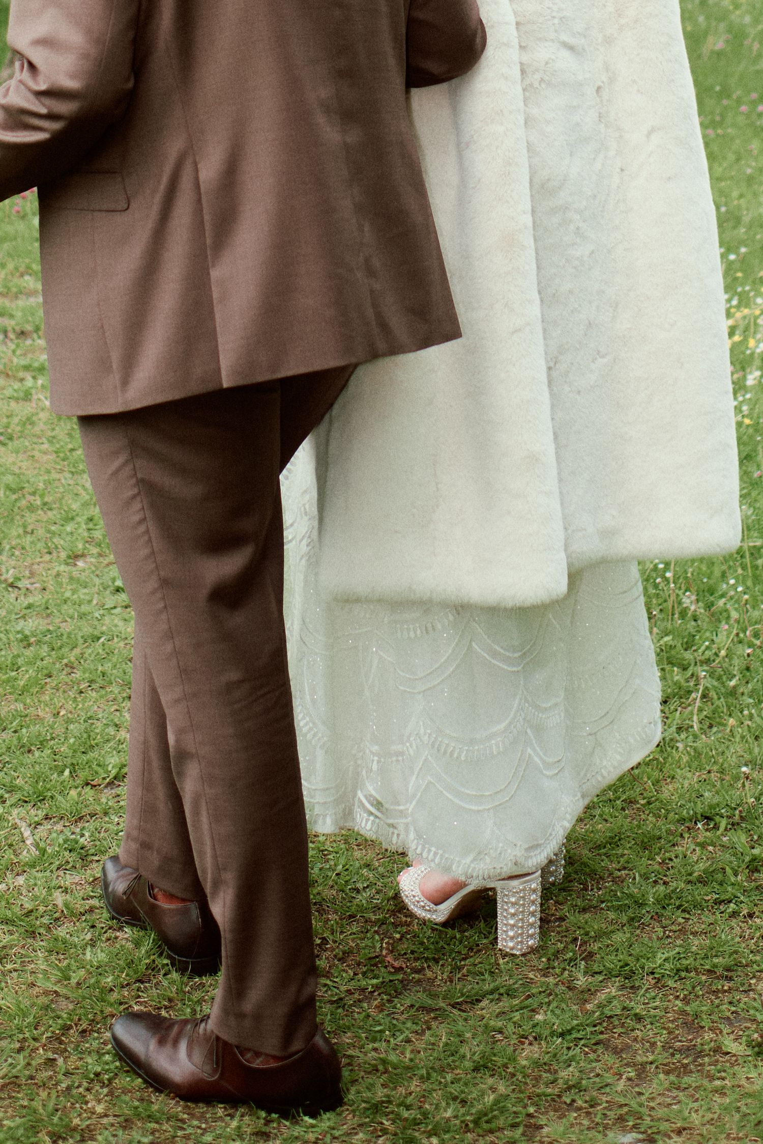 Bride in white lace gown and embellished heels walks beside groom in brown suit on green grass.