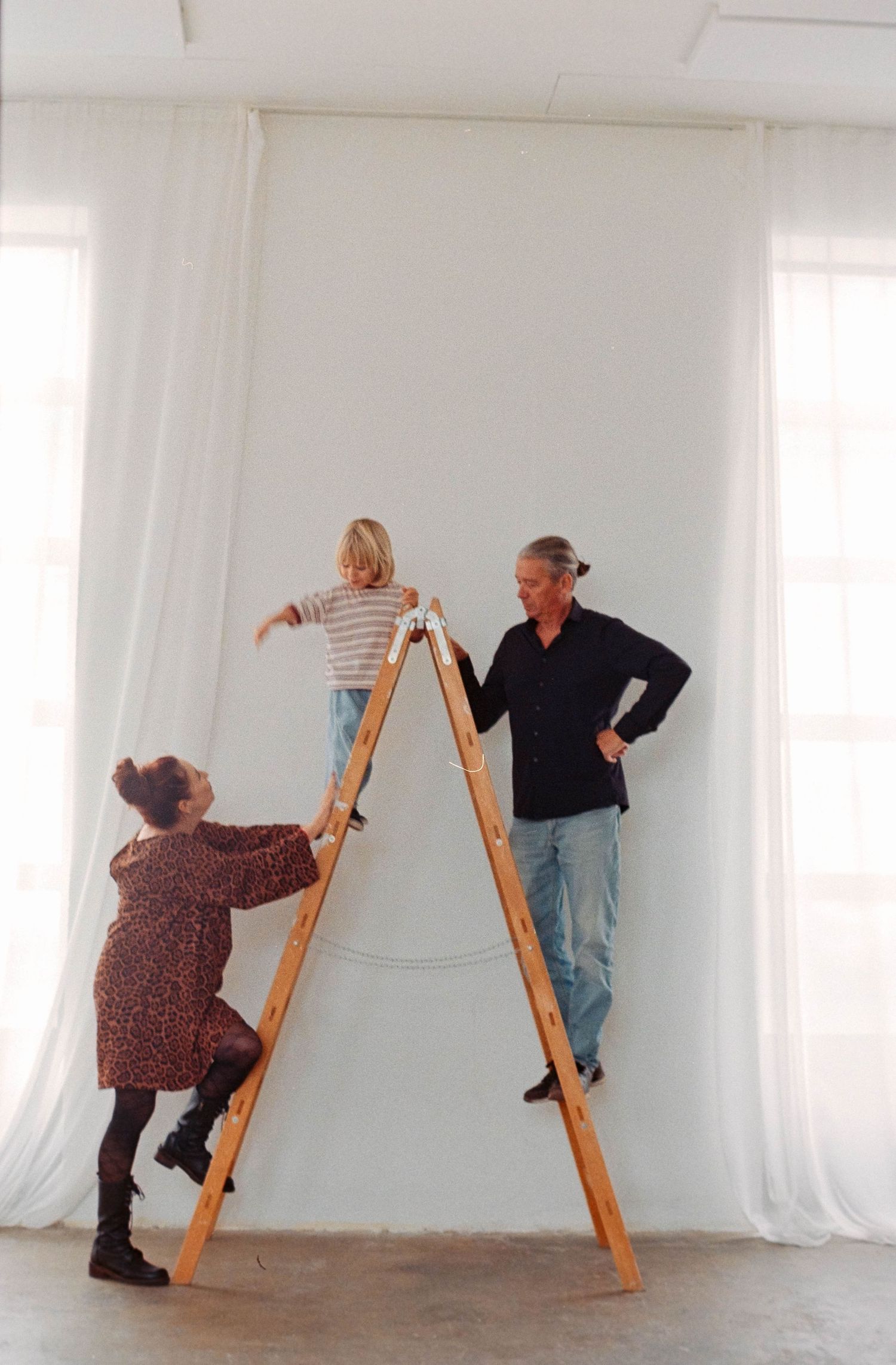 Two adults and a child on a wooden stepladder in a bright white studio space.