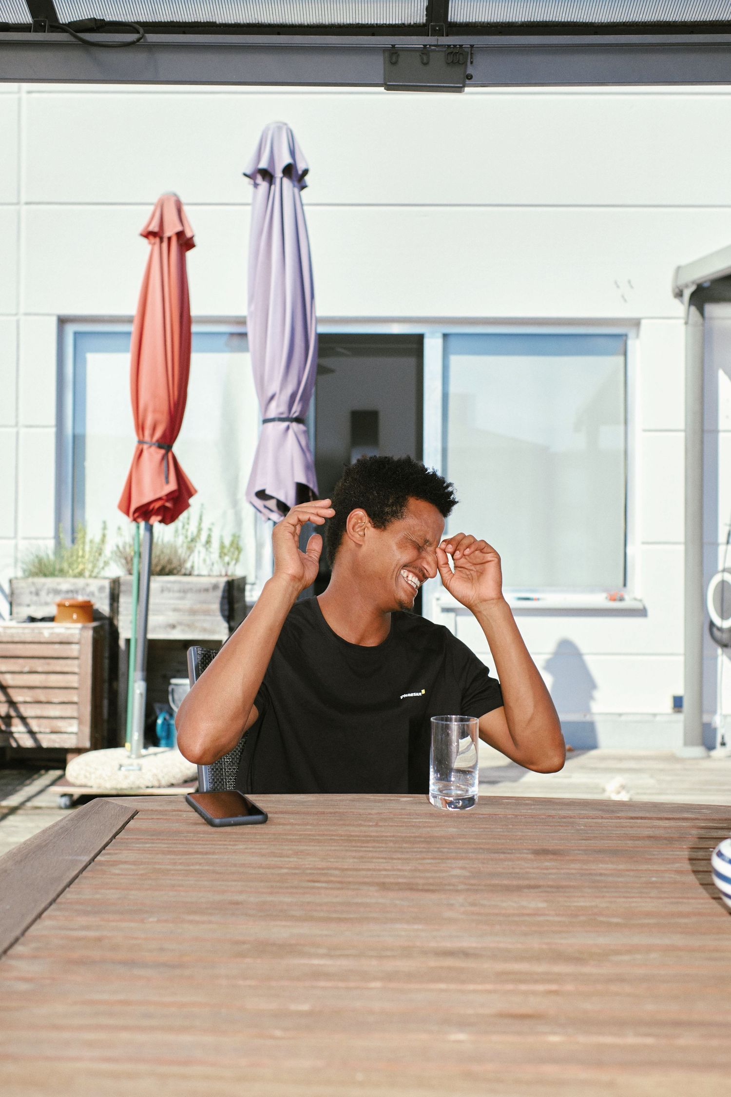 A young man in a black t-shirt laughs while sitting at an outdoor wooden table with a glass of water and smartphone.