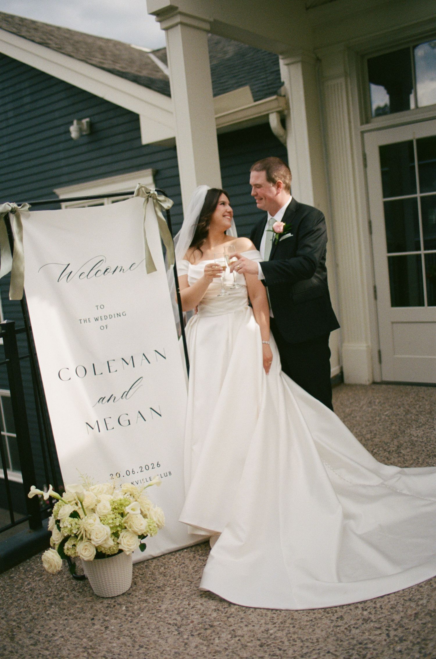 Bride and groom toast beside a welcome sign for Coleman and Megan's wedding outdoors at a venue.