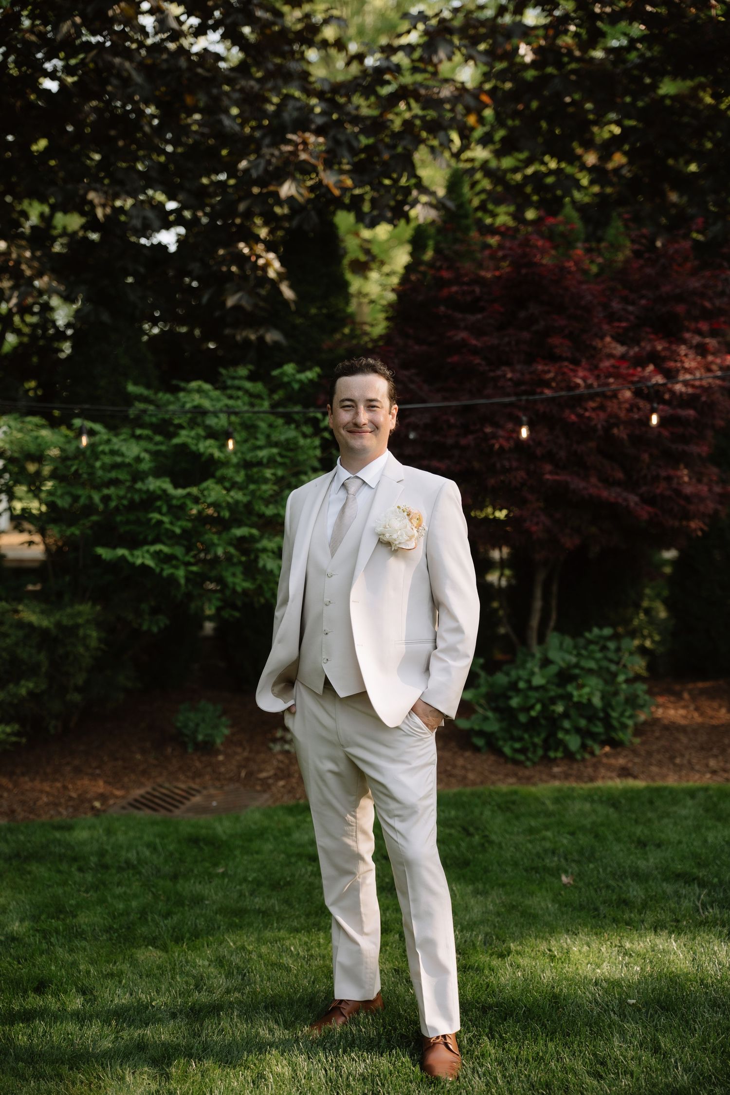 Dapper groom in cream three-piece suit with boutonniere standing confidently on green lawn outdoors.