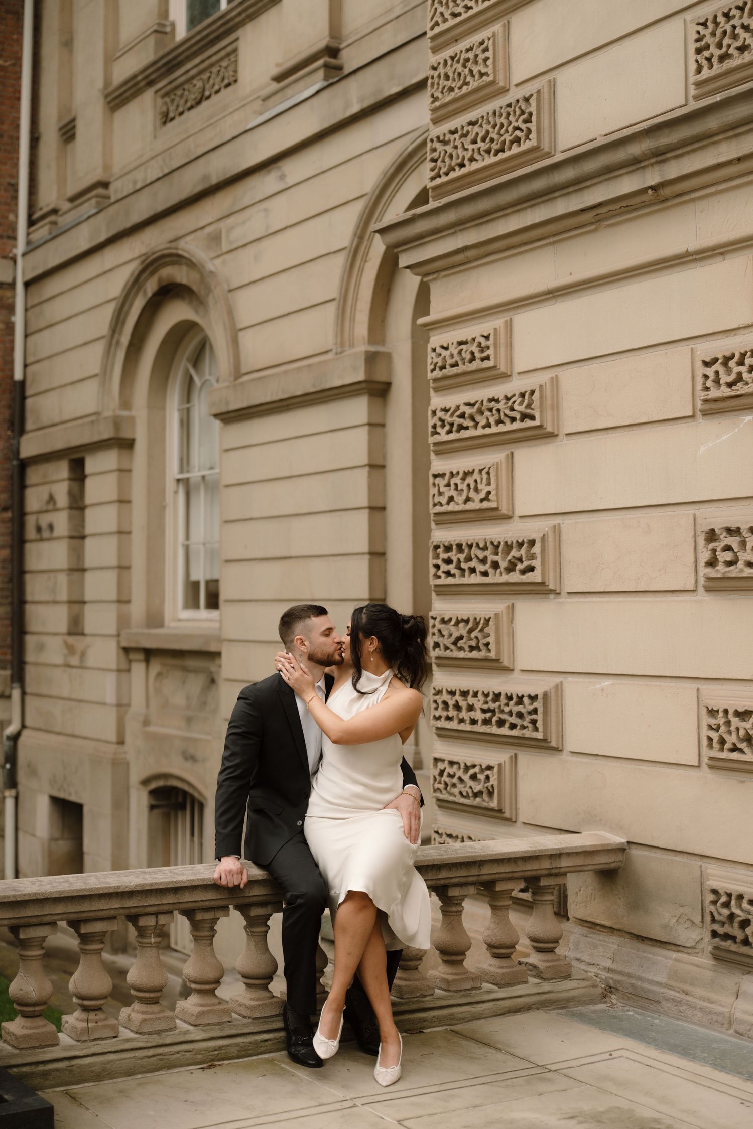 Bride and groom sharing a kiss while seated on ornate stone balustrade of a classic building exterior.