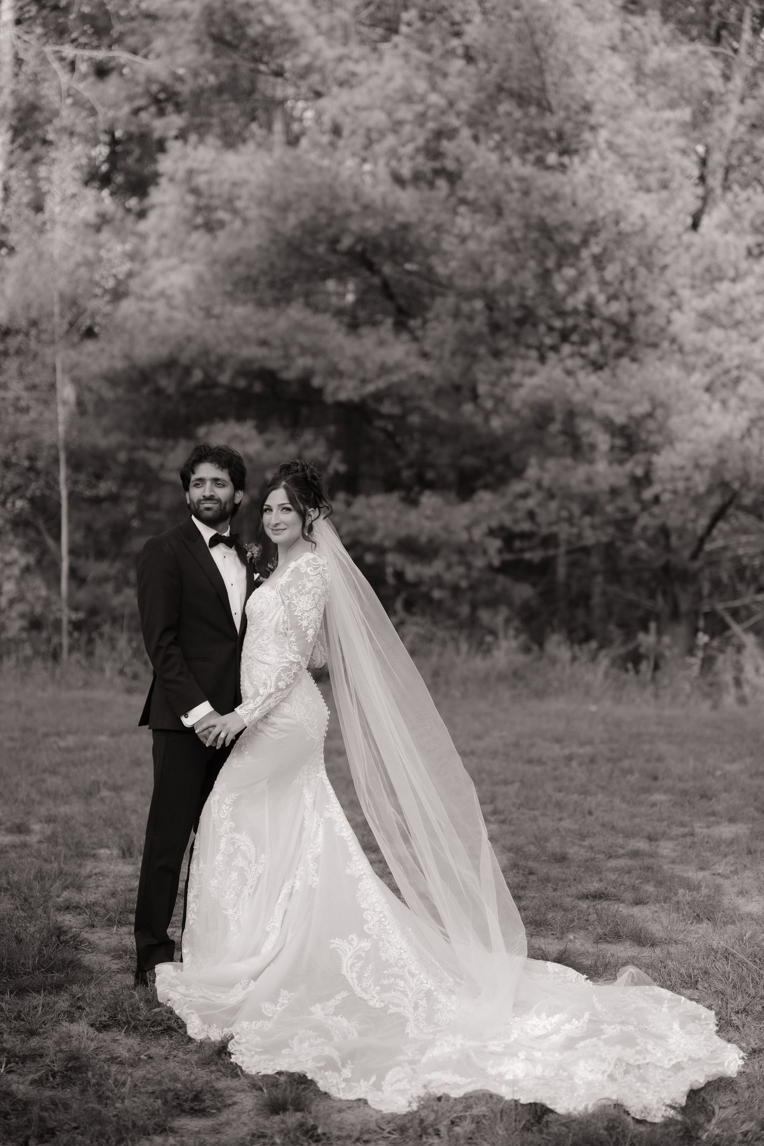 Black and white portrait of bride and groom holding hands in open field surrounded by trees.