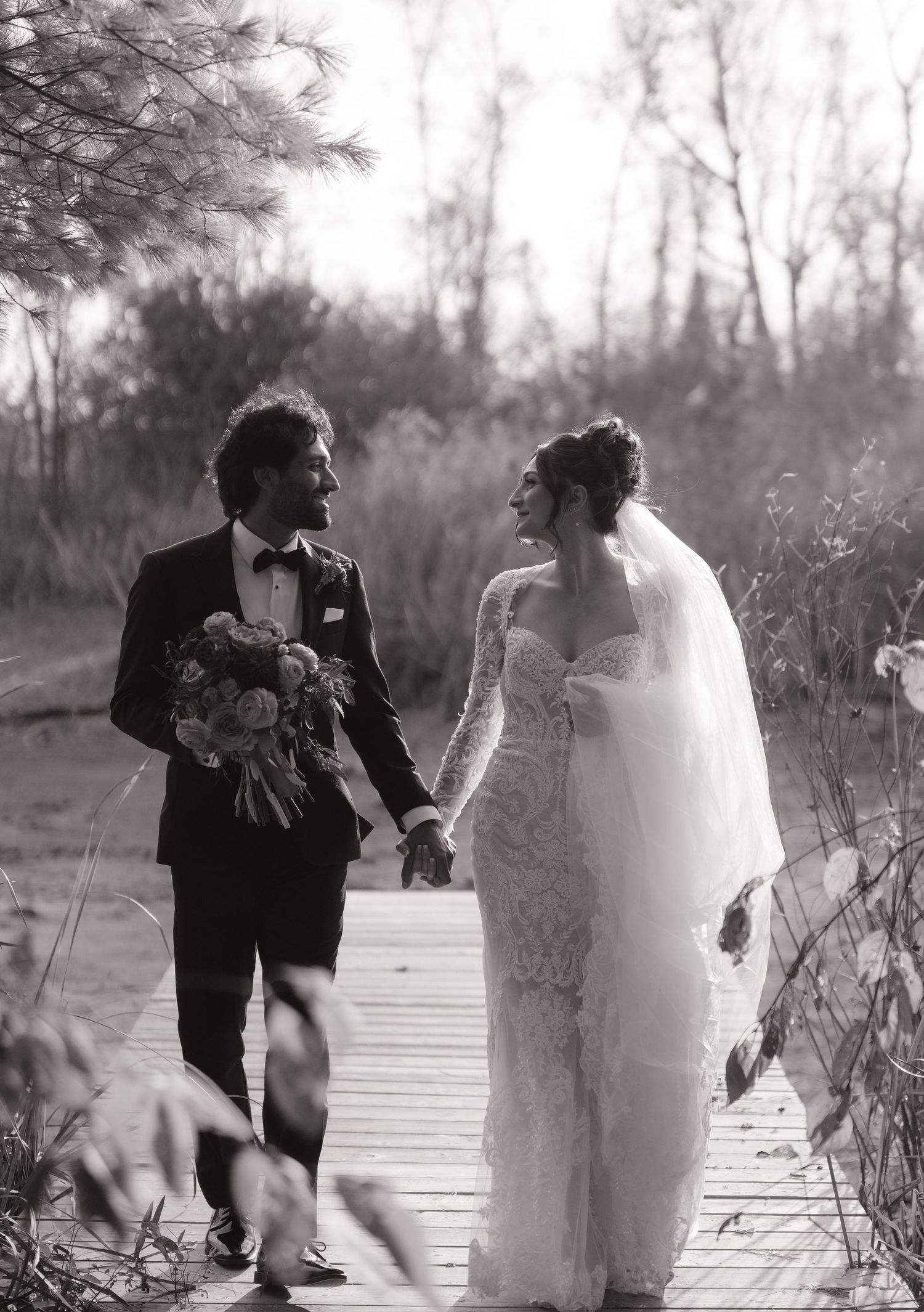 Black and white photo of bride and groom holding hands on wooden boardwalk surrounded by nature.