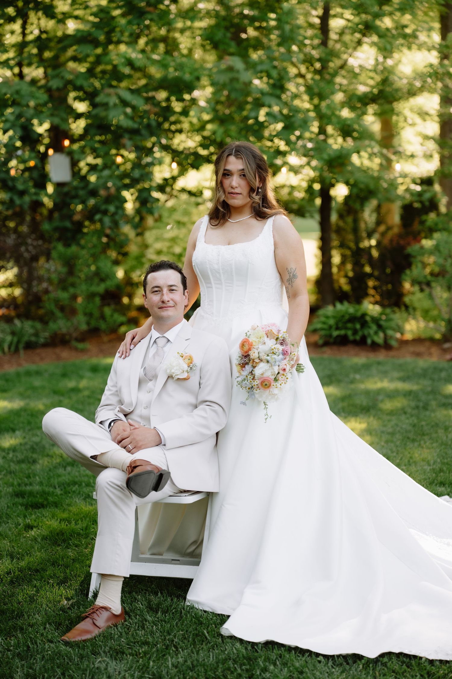 Bride standing behind seated groom in white suit, posing in lush green garden with floral bouquet.