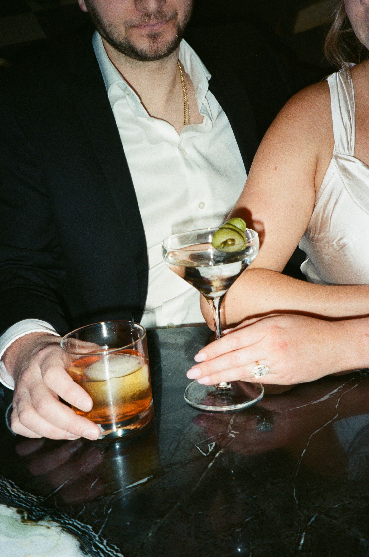 Close-up of engaged couple holding cocktails at marble bar, woman displaying diamond engagement ring.