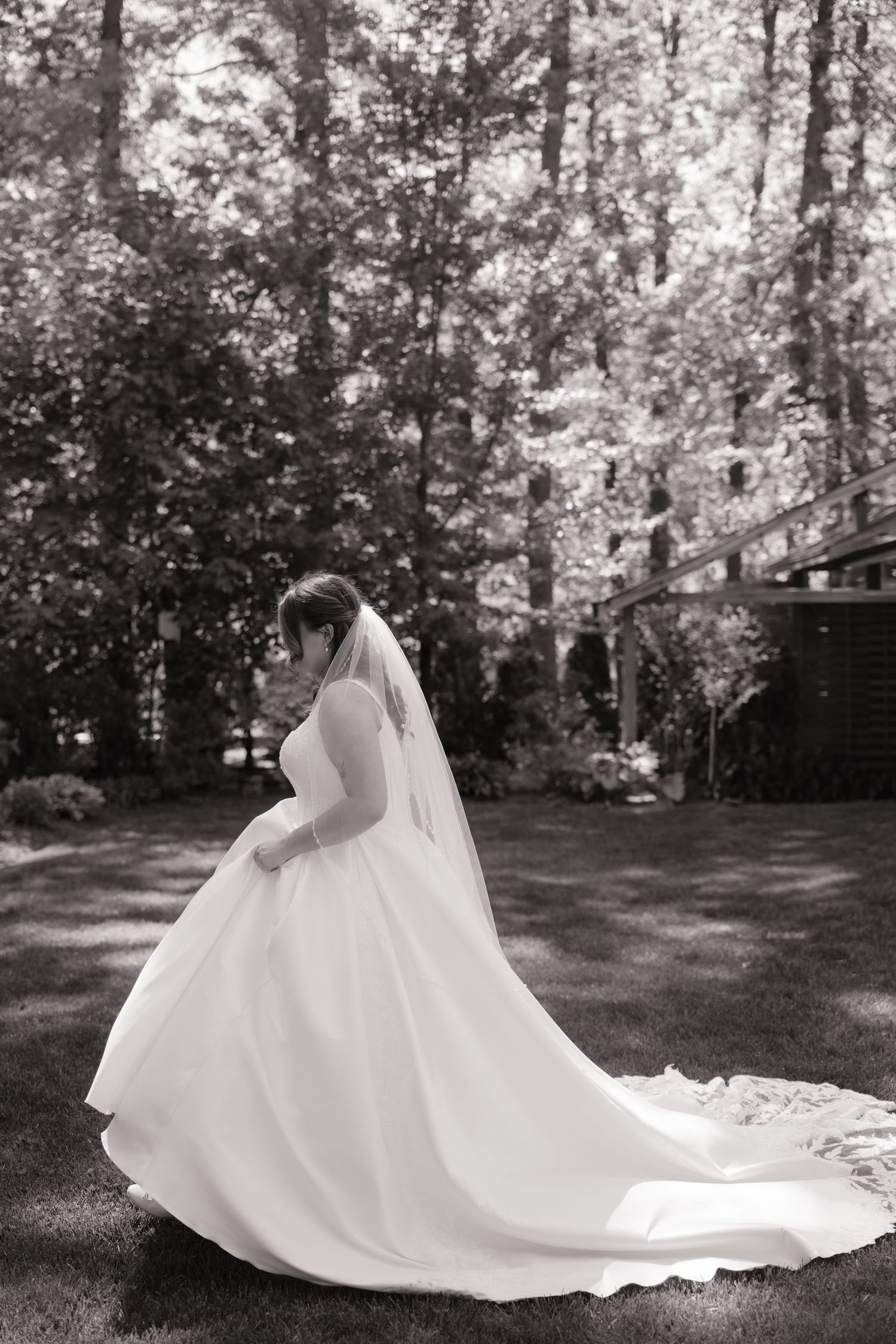 Black and white portrait of bride in ball gown with long veil and train in wooded garden setting.
