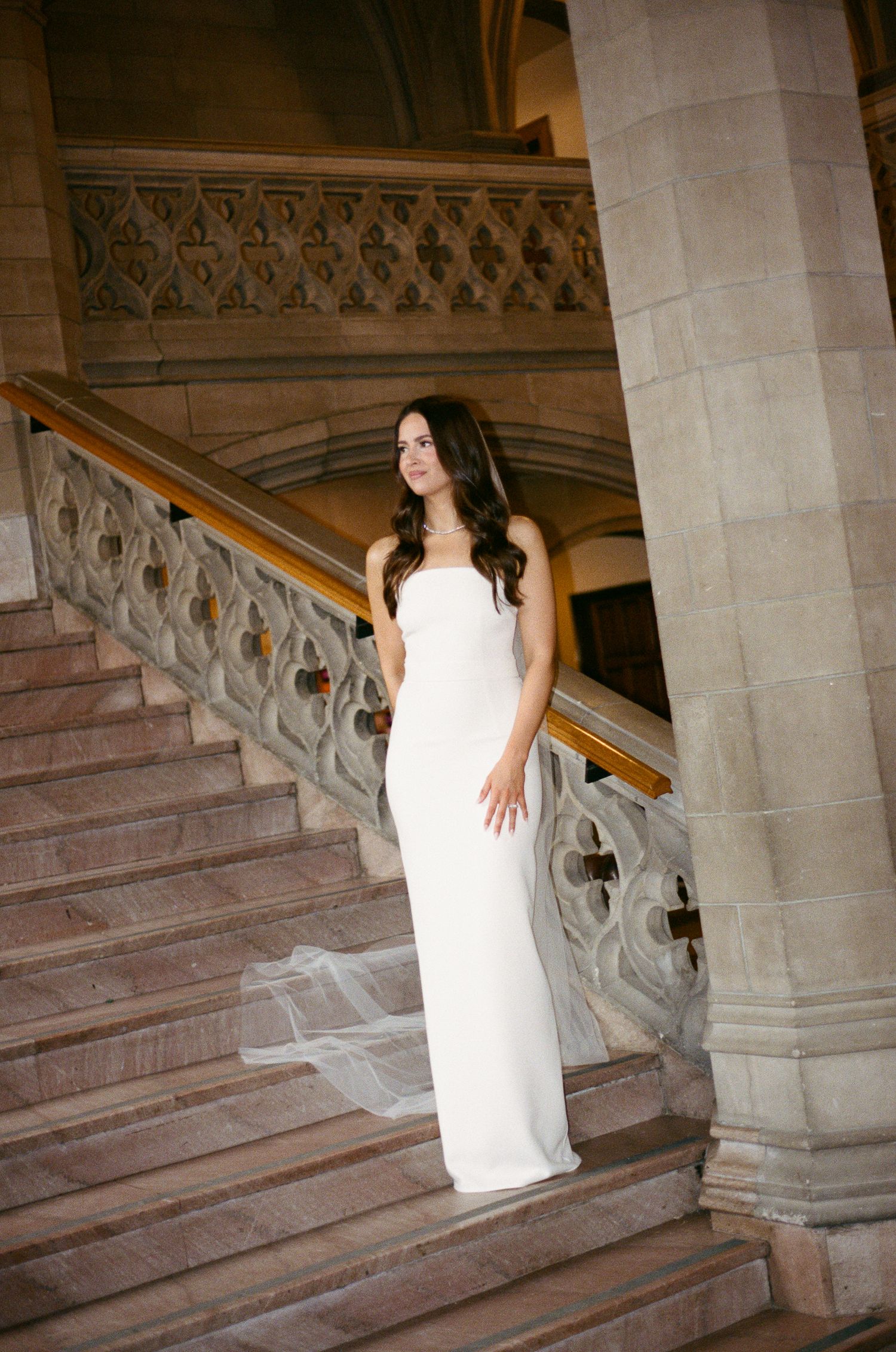 Elegant bride in strapless white column gown with veil posing on grand stone staircase of historic building.