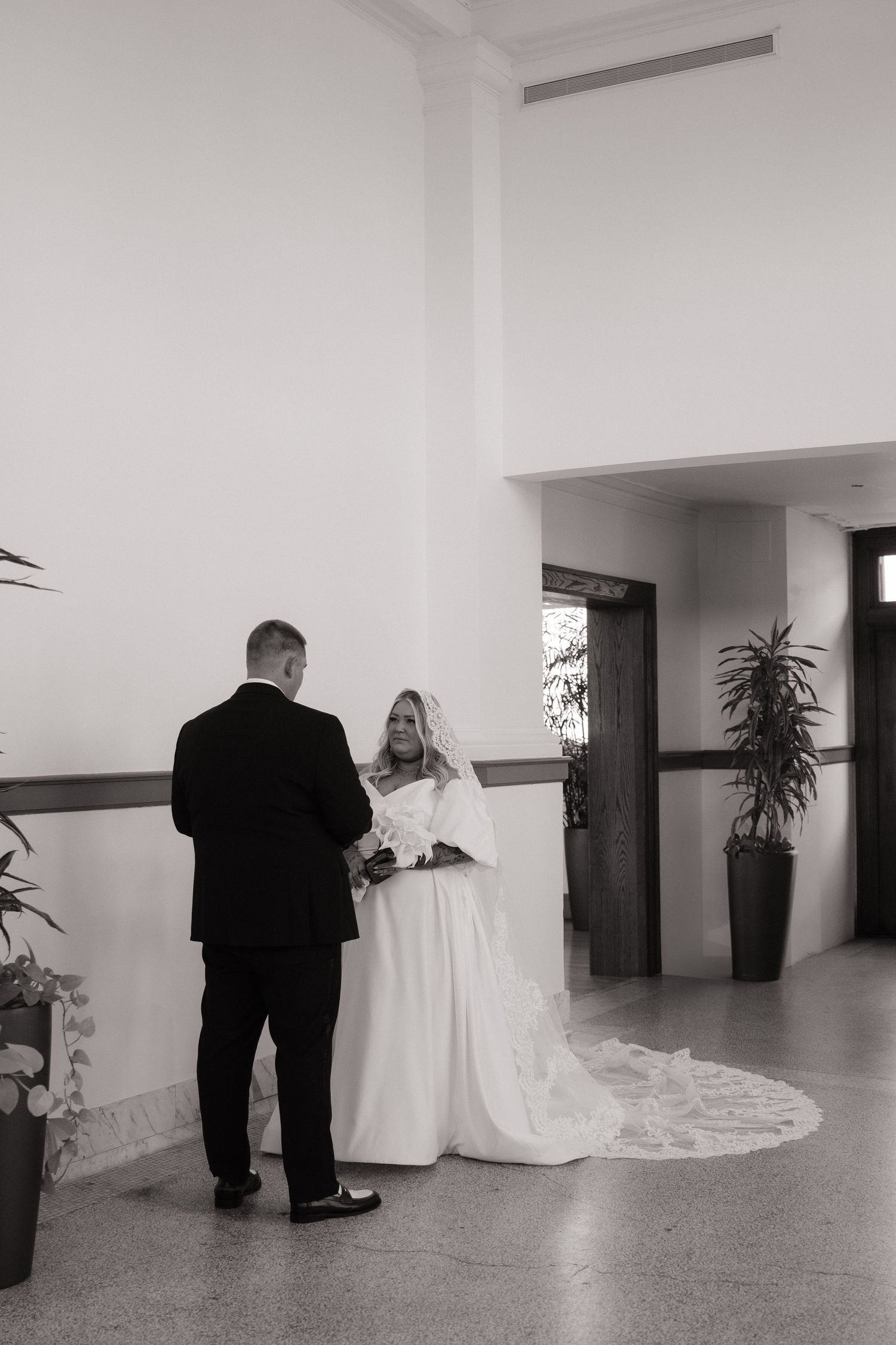 Black and white photo of bride and groom exchanging vows in bright white indoor ceremony space with plants.