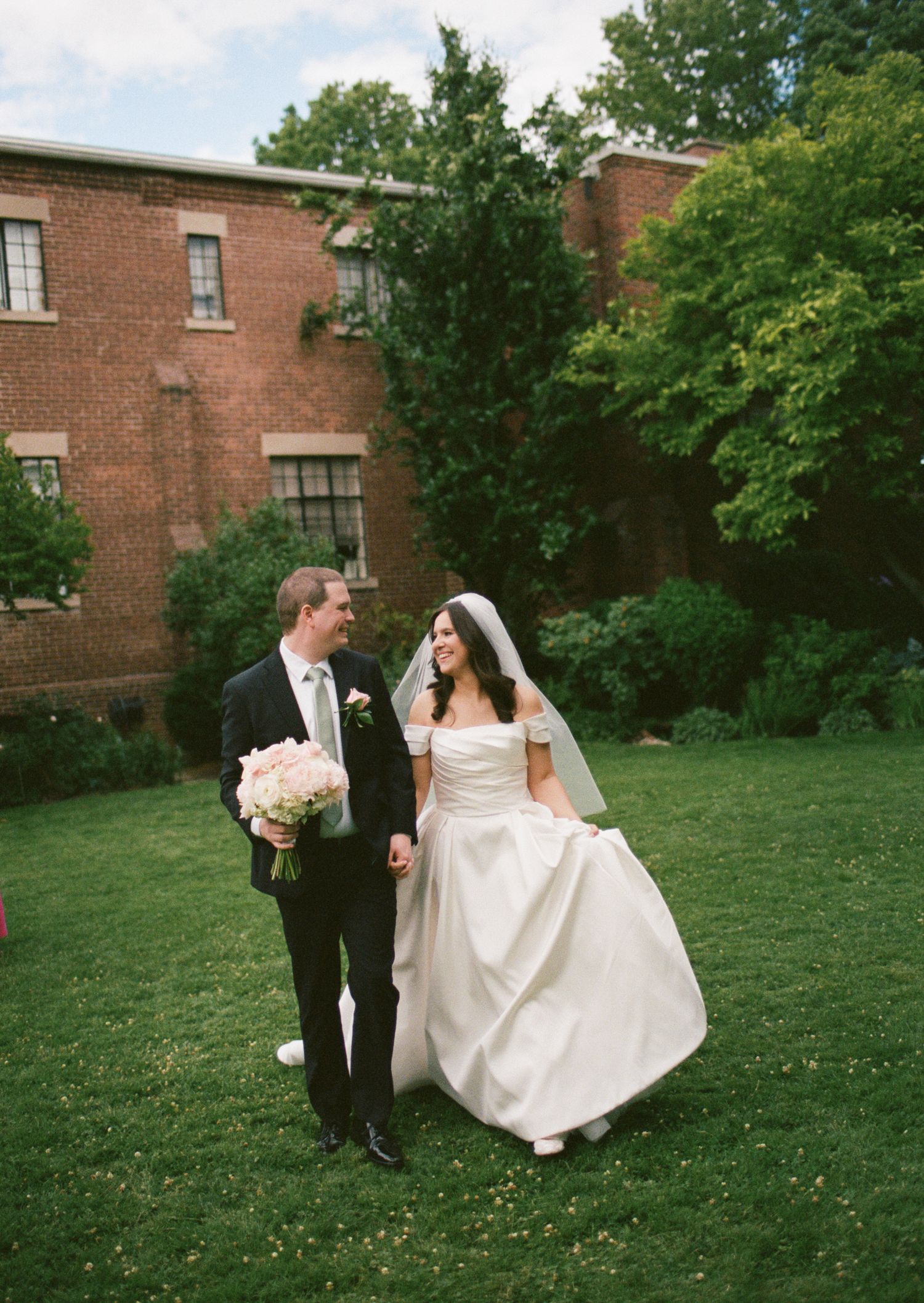 Bride and groom walking hand in hand on green lawn outside brick building, bride holding pink bouquet.